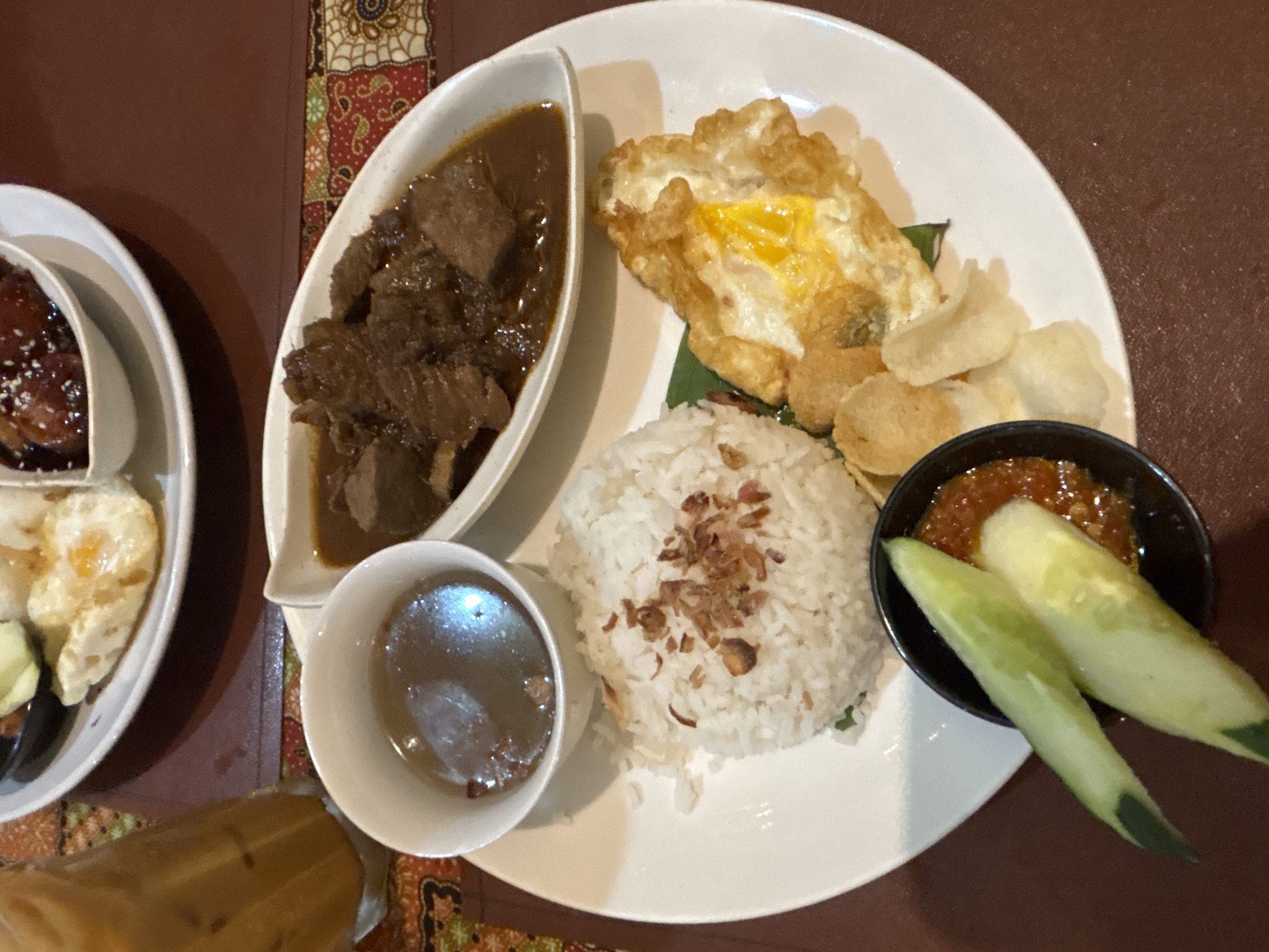 A top-down, close-up shot captures a plate of food at a restaurant in George Town, Malaysia. The dish features a mound of white rice, topped with fried shallots. Adjacent to the rice is a fried egg with a runny yolk and a dark sauce containing bite-sized pieces of meat. A small bowl of clear broth is also present. In the foreground, a small black bowl holds sambal, accompanied by cucumber slices. A few light-colored crackers are scattered on the plate. A tall glass of iced tea sits to the left of the plate. The table is adorned with a patterned placemat. The lighting is dim, suggesting an indoor setting, possibly a restaurant.
