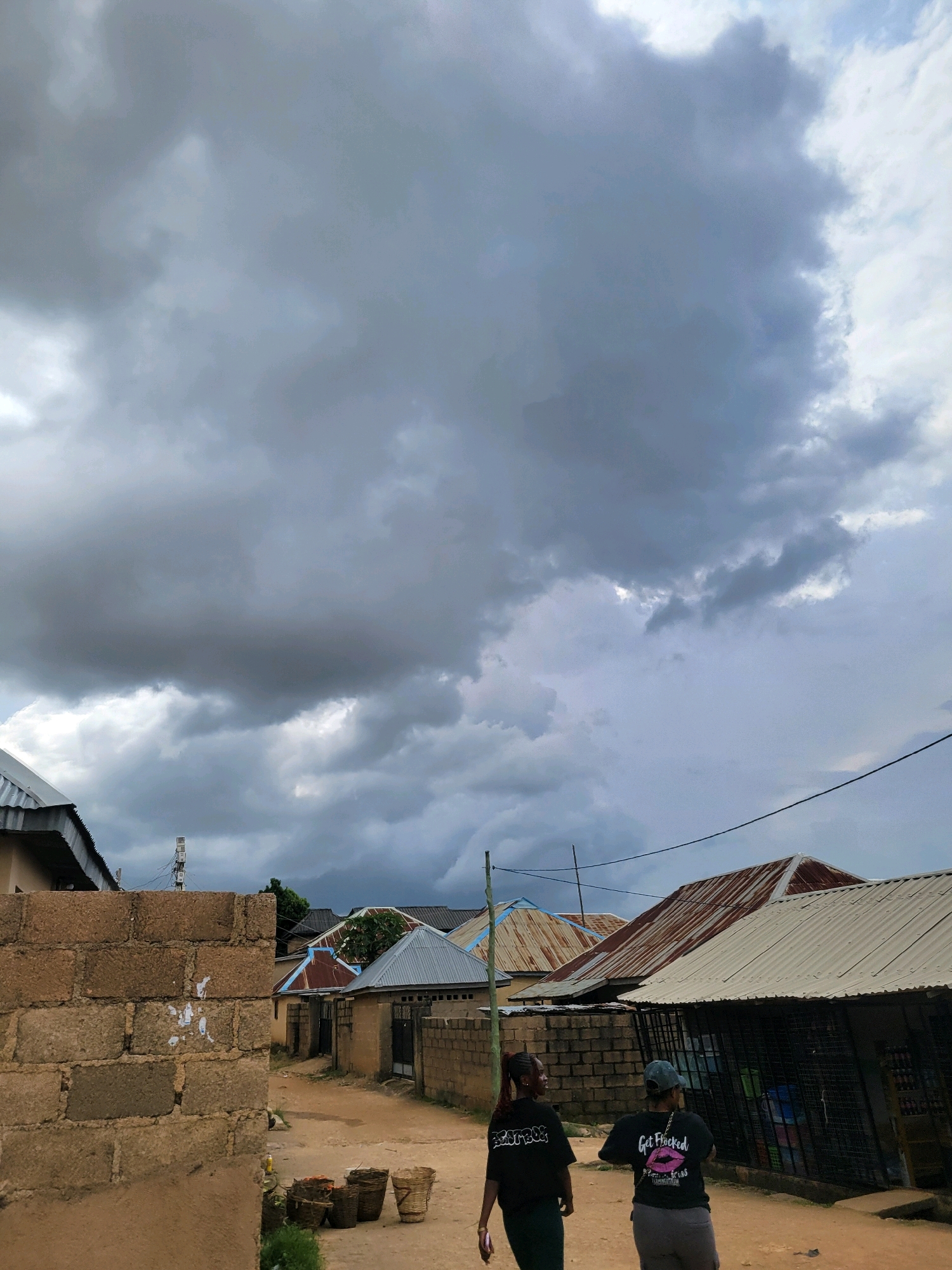 Two women walk down a dirt road in Jos, Nigeria, under a cloudy sky. The woman on the left wears a black t-shirt with "BEATSBO" printed on it and dark pants. The woman on the right wears a black t-shirt with "GET FLOCKED" and a pink lipstick print, and grey pants. They are walking away from the camera towards a row of buildings with corrugated metal roofs. To the left of the road, there is a partially constructed brick wall. Several woven baskets, some containing produce, are visible near the wall. A communication tower is visible in the distance.