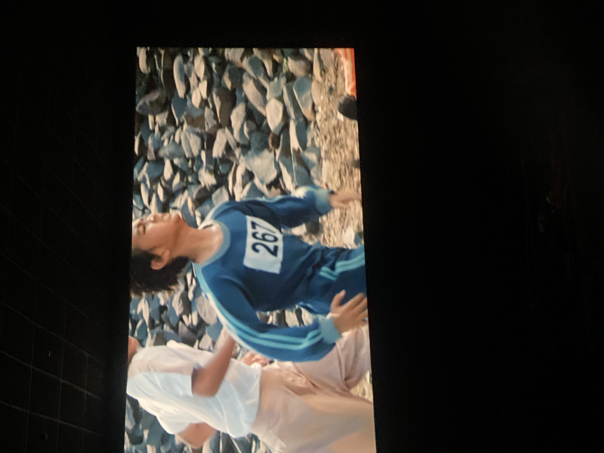 The image shows two individuals in athletic attire running outdoors, with a rocky terrain in the background. The person in the foreground, wearing a blue tracksuit with white and blue stripes, has the number "267" on their chest. The person behind them is wearing a white t-shirt and light-colored pants. The setting appears to be a daytime outdoor scene, possibly a race or athletic training event. There are no discernible elements within the image that would indicate the specific location in Banjarbaru, Indonesia.