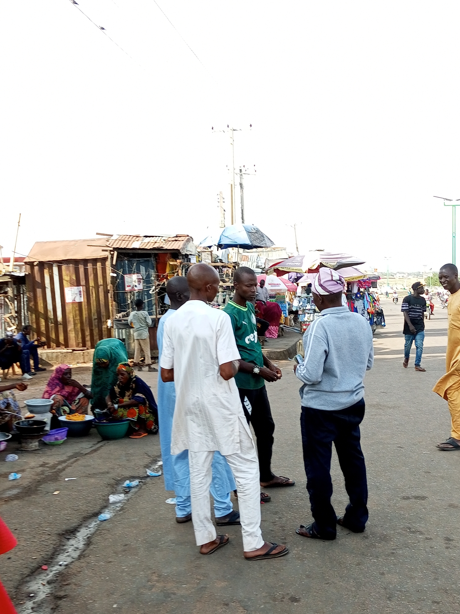This image captures a bustling street scene, likely in Gombe, Nigeria, during the daytime. The overall atmosphere suggests a lively market or roadside gathering.
The primary subjects are a group of men, appearing to be engaged in conversation or some form of social interaction. One man in the foreground wears a white traditional outfit, another a green shirt and black pants, and a third a gray hooded sweatshirt and dark pants. Behind them, other individuals are visible, some tending to stalls or participating in daily activities.
In the background, various structures indicative of a market are present, including corrugated metal stalls and temporary coverings or umbrellas that provide shade. Objects such as bowls and what appear to be food items are visible in the lower left, suggesting a food vendor or a place where goods are being sold or prepared. The road surface is asphalt, and there are some debris and water puddles, hinting at recent rain or poor drainage.
The sky is overcast and bright, suggesting it might be a slightly hazy or cloudy day, common in many parts of Nigeria. The lighting is even, without harsh shadows, further supporting a daytime setting. There are no discernible emotions of distress or extreme joy, the general mood seems to be one of everyday commerce and community interaction.
No visible text is clearly legible in the image to provide specific location details or identify any businesses. The presence of the stalls, the clothing styles of the people, and the general ambiance strongly point towards a Nigerian setting, specifically Gombe as indicated by the context. The scene depicts a slice of daily life, emphasizing the vibrant social and economic activities typical of a Nigerian street market.