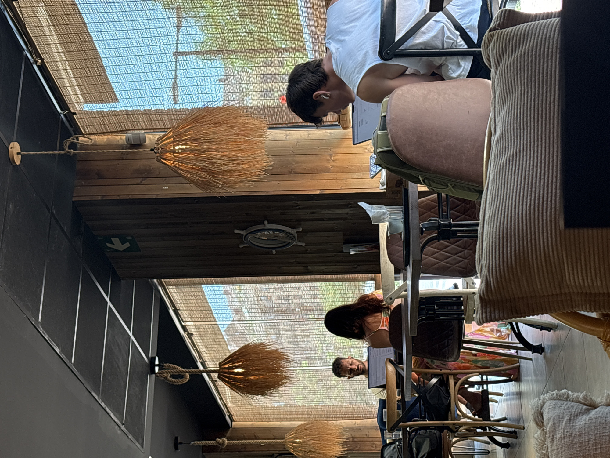 This image shows the interior of a cafe or restaurant, likely in València, Spain, with a focus on people seated at tables and the surrounding decor. The setting appears to be a space with a sloped ceiling covered in bamboo blinds, allowing natural light to filter through. Wooden panels line the walls and ceiling, creating a warm and rustic ambiance.
In the foreground, a person is seen from the back, seated at a table with a laptop. Further into the scene, another person, a young man, is bent over a laptop, wearing earbuds. He is wearing a white t-shirt and appears focused on his work. To his right, a person with long, dark hair, possibly a woman, is also seated, looking towards a screen. Another man with a beard is visible in the background, also looking down.
The cafe has unique lighting fixtures made of straw or similar material, resembling brooms, suspended from ropes. The furniture includes various chairs, some with brown padded seats and backs, and others made of lighter wood. The overall atmosphere suggests a relaxed daytime setting, possibly late morning or early afternoon, given the bright natural light. The presence of people working on laptops and enjoying the space indicates it's a functional cafe environment.