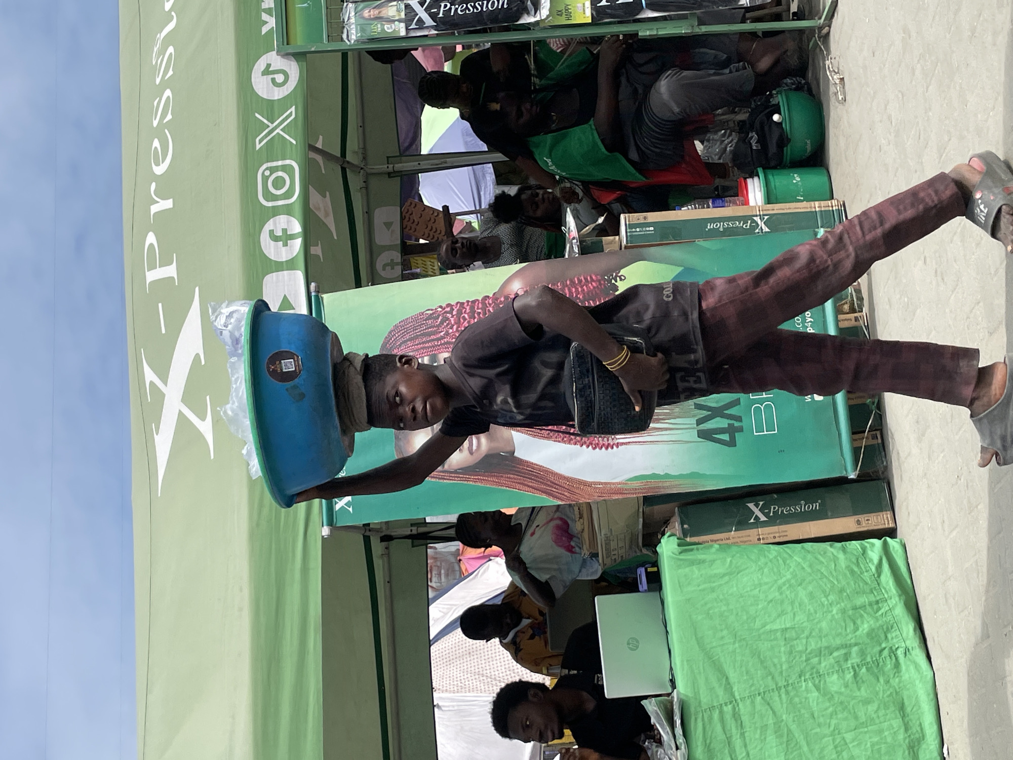 A young boy is walking in a market in Lekki, Nigeria, carrying a blue bowl filled with plastic bags on his head and a black box in his right hand. He is wearing a dark t-shirt and checkered pants. In the background, a green tent with the logo "X-Pression" is visible, along with several people. Some people are seated, while others are standing. There are also some products displayed, including what appear to be hair extensions. The scene suggests a busy marketplace environment.