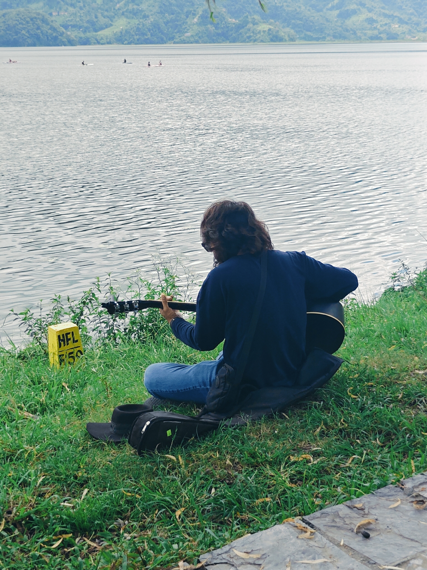 A person with long brown hair is playing an acoustic guitar while sitting on the grassy bank of Phewa Lake in Pokhara, Nepal. The person is wearing a dark blue long-sleeved shirt and blue jeans. In front of the person, on the grass, are a black bag and a black hat. To the left of the person, a yellow marker post with "HFL 150" written on it is visible. The lake stretches out behind the person, with distant green hills and mountains in the background. Several people are visible on paddleboards in the distance on the lake. The water is rippled and reflects the sky. The overall scene depicts a serene, outdoor setting.