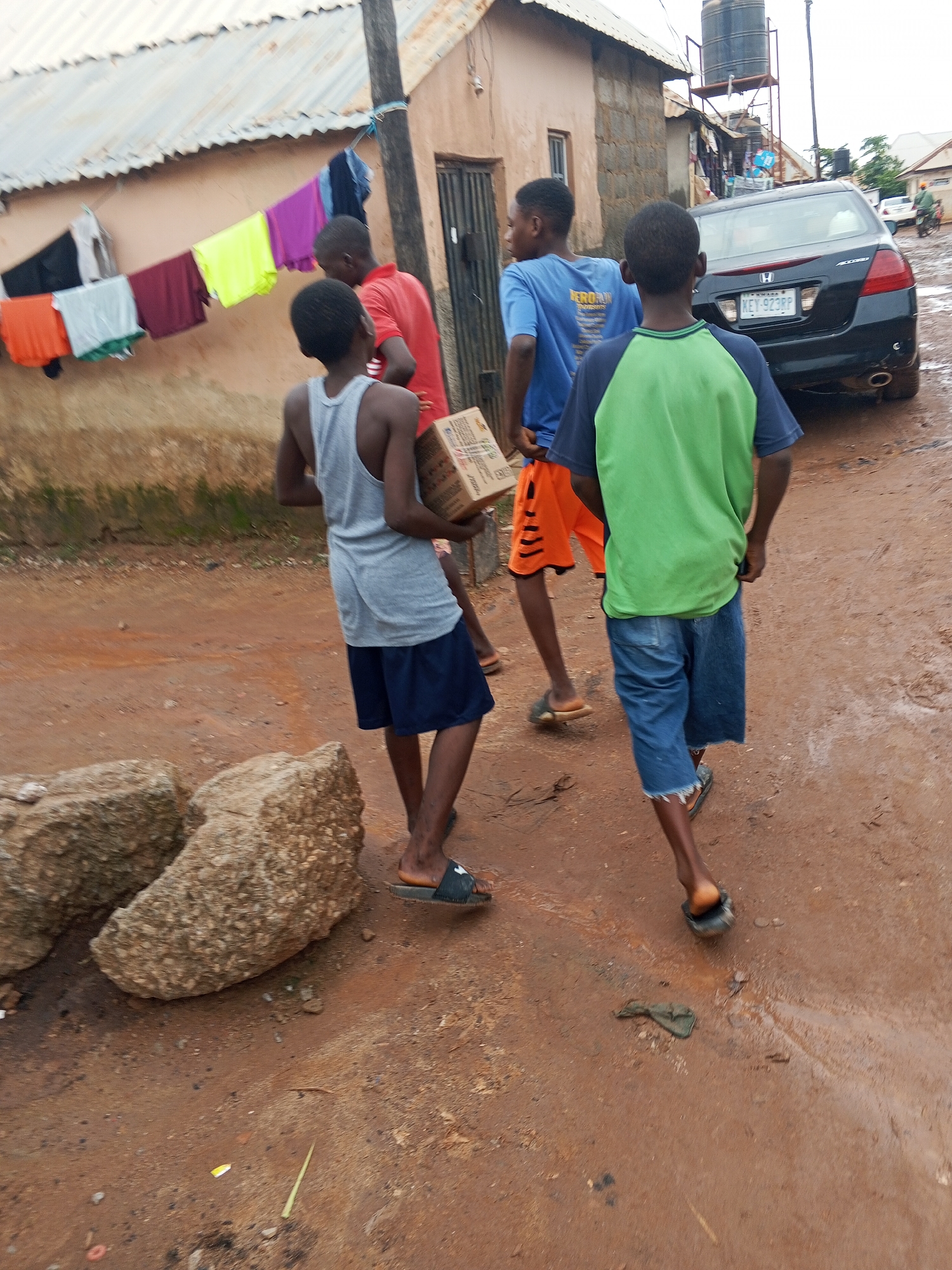 The image shows three young boys walking on a dirt road in Abuja, Nigeria. The road appears to be wet, suggesting recent rain. The boys are dressed casually, with one wearing a grey tank top and dark blue shorts, another in a red t-shirt, and the third in a blue t-shirt and orange shorts. The boy in the grey tank top is carrying a cardboard box. Behind them, a black Honda Accord is parked, and a motorcycle is visible in the distance. Clothes are hanging on a line to dry against a building with a corrugated iron roof. The scene suggests a residential or semi-rural area. The overall mood seems ordinary, with the boys engaged in everyday activity. The weather appears overcast. There is no visible text in the image.