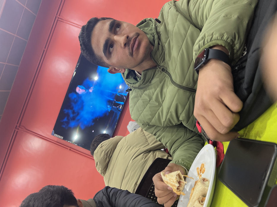 The scene depicts the interior of a casual eatery in Rukn ad-Din Municipality, Syria. A young adult male, with short dark hair and a light mustache, is positioned in the foreground, seated at a lime-green tabletop. He wears an olive-green quilted puffer jacket and a dark smartwatch on his left wrist. His gaze is directed towards the right side of the frame, away from the camera. His right hand holds a partially consumed wrap sandwich, revealing a light-colored filling. On a white plate to his left, additional wrap pieces and scattered orange-reddish crispy fragments are visible. A black smartphone lies face down on the table near his left hand, which rests on a dark bag or fabric item.
To the left of the primary subject, the shoulder and partial head of another individual in a dark jacket are visible. Further in the background, a third person is seated with their back to the camera, wearing a light tan or beige quilted puffer jacket. This individual is seated in a chair with a dark, perforated backrest. The background wall is painted a vibrant red, featuring horizontal and diagonal light-colored piping. A large flat-screen television is mounted on this wall, displaying an image with intense blue illumination surrounding a dark, humanoid figure. The overall lighting is artificial.