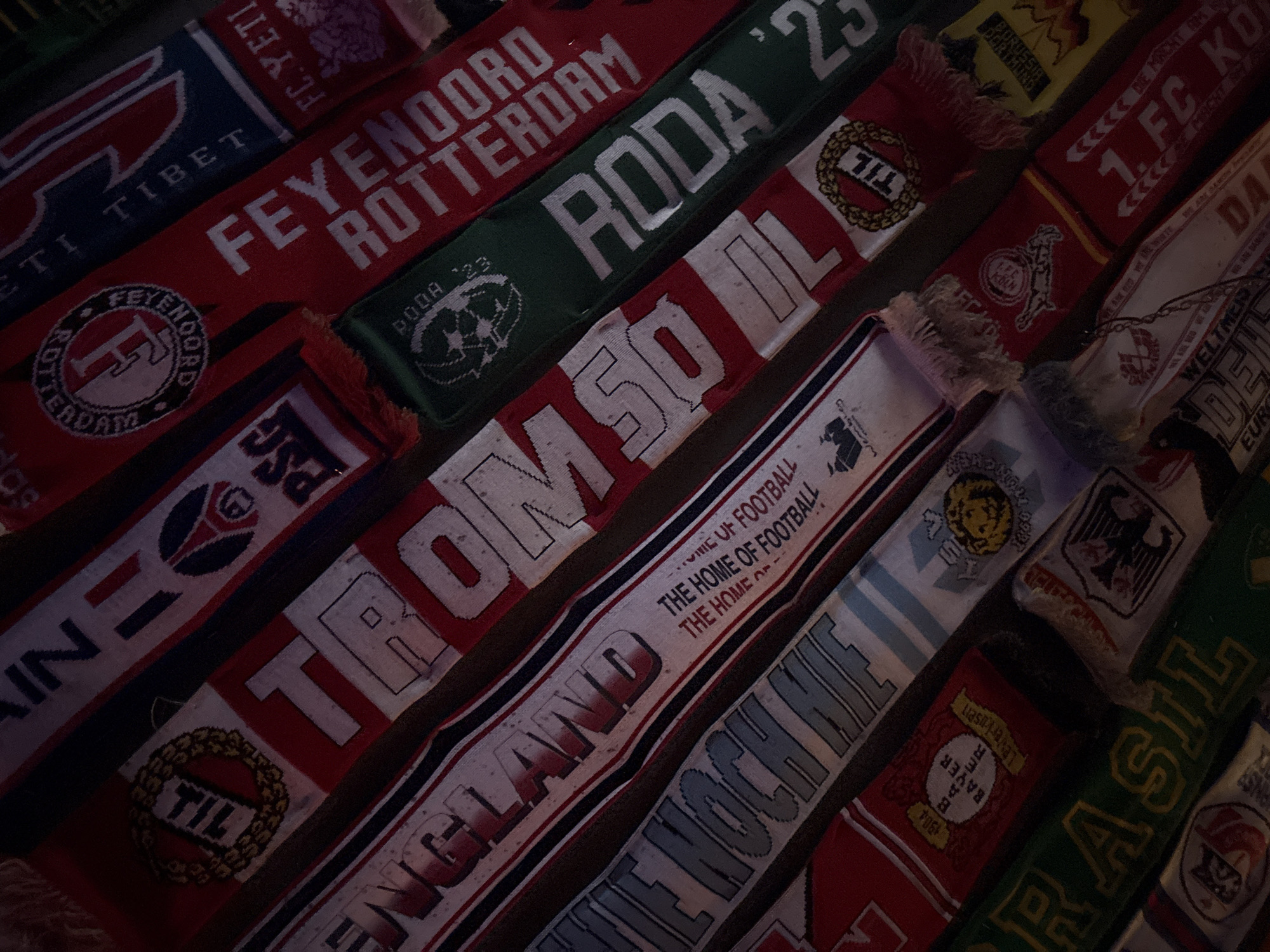 The image displays a collection of football scarves, each representing a different team or country. Prominent teams/locations visible include Feyenoord Rotterdam, Roda JC Kerkrade, Tromsø IL, Paris Saint-Germain (PSG), England (with the text "The Home of Football"), 1. FC Köln, Bayer Leverkusen, and Brazil. There is also a scarf with "FC YETI" written on it, and another for "Tibet." The scarves are arranged in a slightly overlapping pattern, filling the frame. The lighting is dim, and the image appears to be a close-up. There is no information in the image to associate with Plovdiv, Bulgaria.