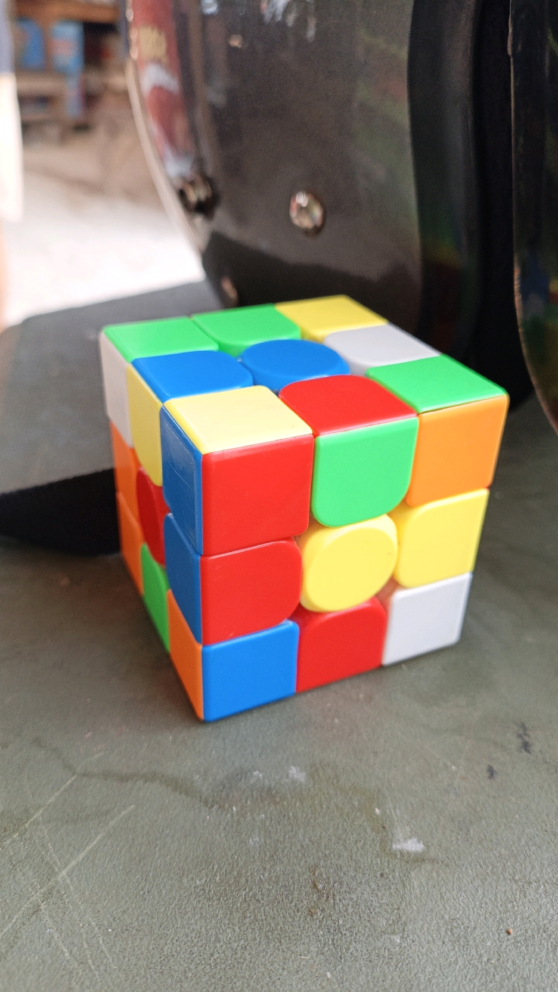 A cube puzzle sits on a textured, green surface. The puzzle is multicolored with red, blue, green, yellow, orange, and white squares. The background is out of focus, showing what appears to be a large, dark, curved object, possibly a motorcycle helmet, and a blurry outdoor scene with wooden structures. The specific location in Cikal, Indonesia, is not discernible from the image.