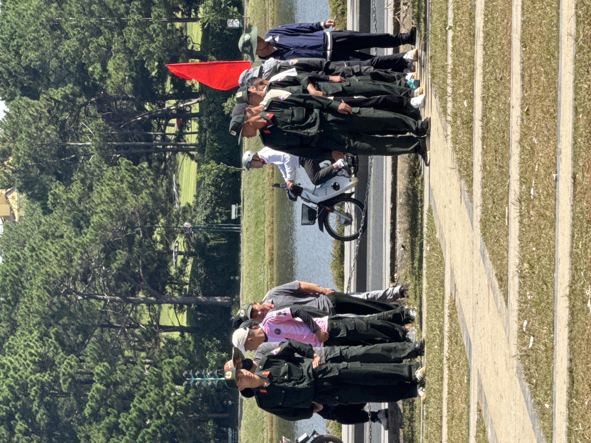 The image depicts a group of men, some in military-style uniforms, standing in what appears to be a park or recreational area in Da Lat, Vietnam. The setting features a body of water, lush green trees resembling pine trees, and a paved pathway with grassy sections. In the background, there's a red flag fluttering on a pole. To the right, a person on a scooter is visible, and further in the distance, more trees and a hint of structures suggest a developed area. The men are positioned in a line, with some looking towards the camera and others looking in different directions, suggesting they might be posing or engaged in some activity. The overall atmosphere is bright and sunny, indicating daytime.