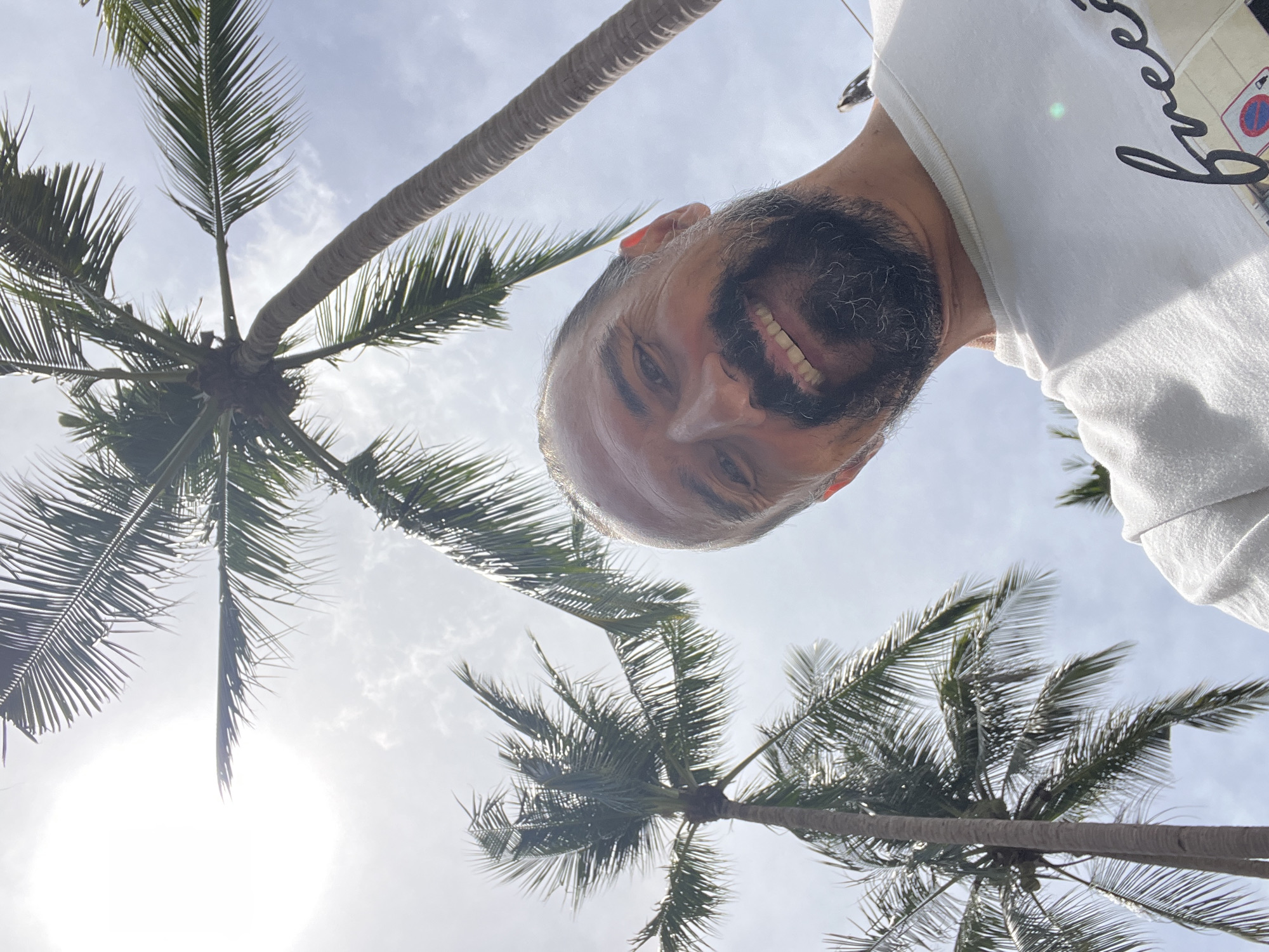 A low-angle, upwards-facing shot shows a smiling man with a beard and gray hair, wearing a white t-shirt with black text, against a backdrop of several tall palm trees silhouetted against a partly cloudy sky. The location is identified as บ้านบางรักษ์, Thailand.