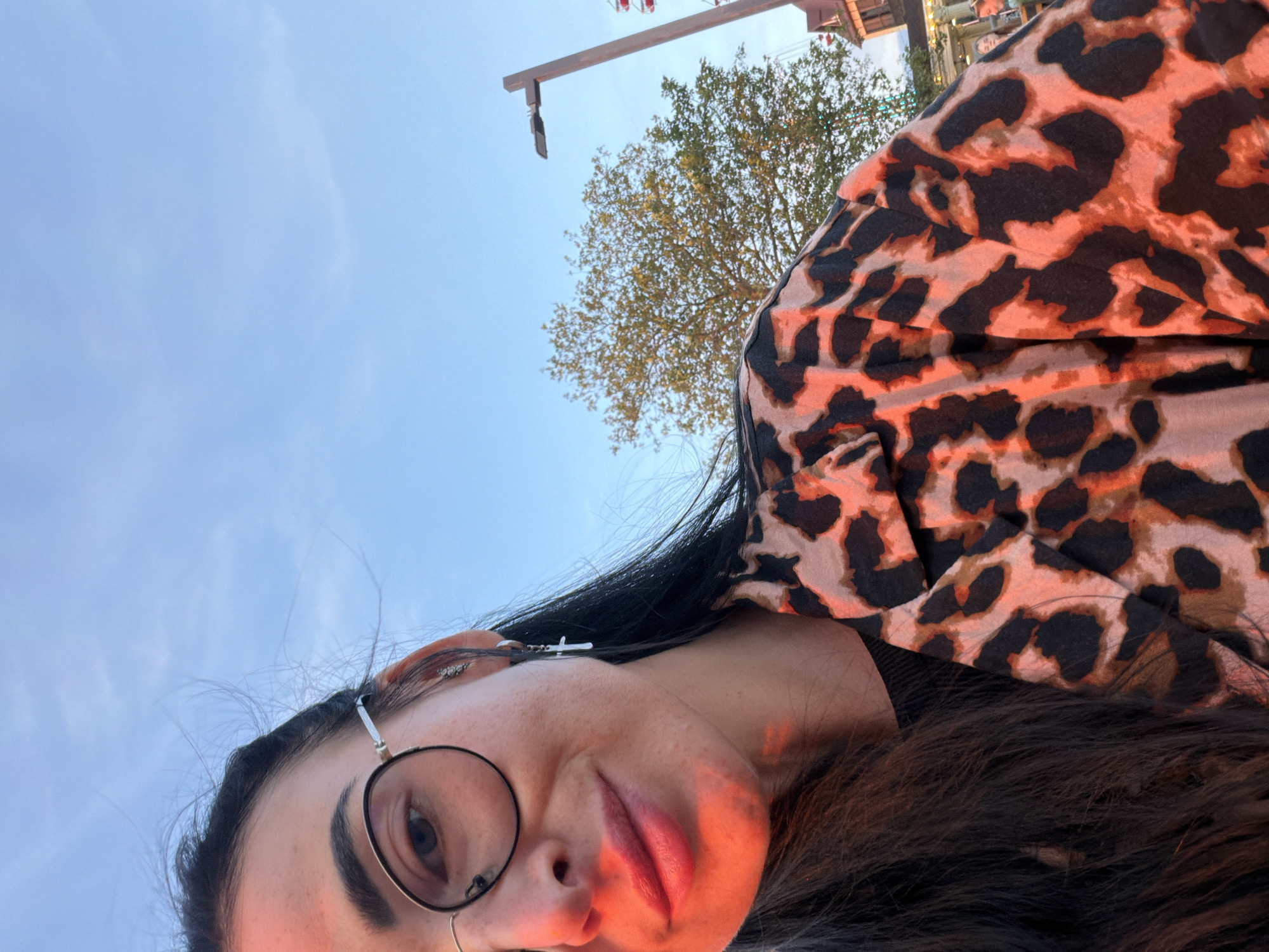 A close-up, medium shot captures a woman with dark hair, wearing round-framed glasses and a leopard-print shirt. Her face, angled towards the camera, is illuminated by a warm, orange light, casting shadows and highlighting her features. She is smiling slightly. Her left ear displays a silver hoop earring with a small cross pendant. The background features a clear blue sky, a light pole with a fixture, a leafy green tree, and hints of a building or structure with red accents. The image appears to be taken outdoors, possibly in a public space like an amusement park, given the presence of a Ferris wheel and other carnival-like structures visible in the distance.