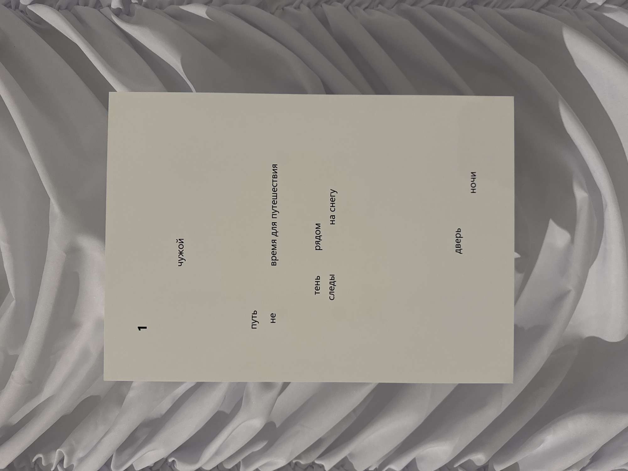 The image depicts a sheet of off-white paper placed against a backdrop of white, gathered fabric, possibly a curtain or draped material. On the paper, text is printed in a simple, dark font. At the top left, the number "1" is prominently displayed. Below this, the word "чужой" is written. Further down, the word "путь" appears on the left, with "время для путешествия" to its right. Below "путь" is the word "не". Towards the bottom left, "тень" is written above "следы". To the right of "тень" is "рядом", and below that, "на снегу". At the bottom center, "дверь" is printed, and to its right, "ночи". The overall composition suggests a minimalist presentation of text, likely an excerpt from a poem or literary work, set against a textured, neutral background. There are no people or specific Moscow landmarks visible in the image.