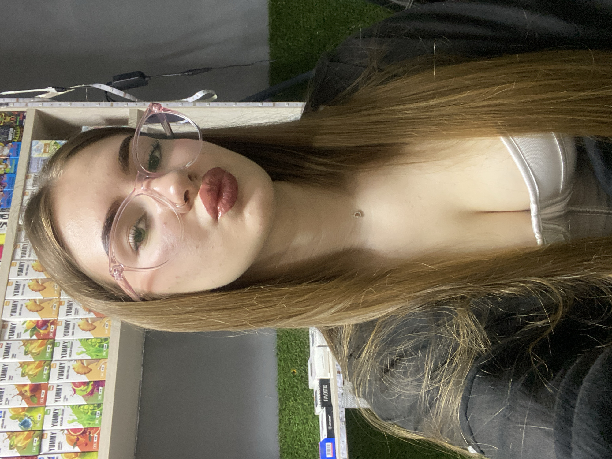 The image shows a young woman with long, light brown hair, wearing pink-rimmed glasses and a dark jacket over a white top. She has a fair complexion and is wearing lip gloss. She is wearing a small, delicate necklace. The background appears to be indoors, with shelves in the background displaying numerous small boxes of products, possibly vape liquids, with colorful graphics. There is a green, artificial grass-like material visible on a lower shelf. A strip of LED lights is attached to a vertical pole on the right side of the frame, and a power adapter is plugged into an outlet on the same pole. The overall impression is of a retail environment, potentially a vape shop. The specific location is in the city of Dyakovo, Russia.