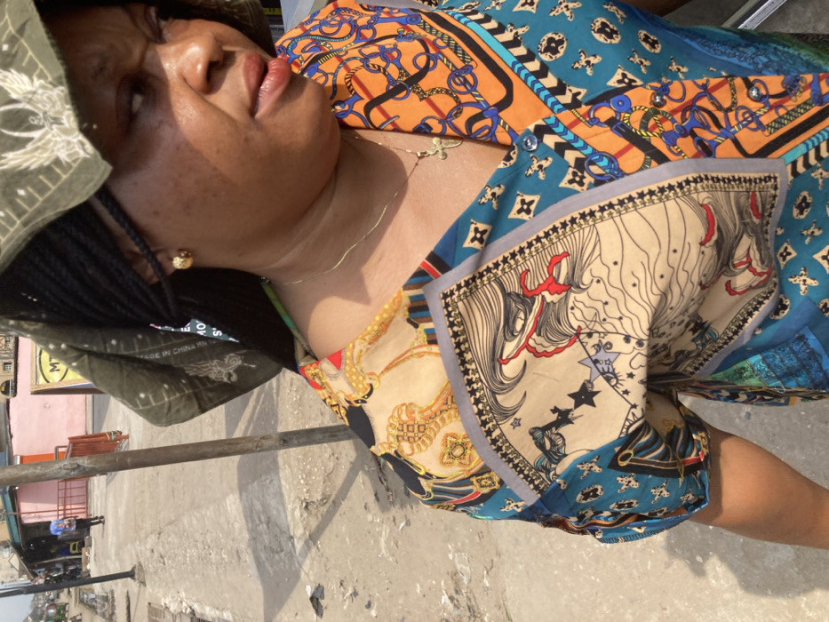 The scene depicts an adult female subject in a bright, outdoor urban environment identified as Umuode, Nigeria. The subject, positioned in the foreground and mid-shot, is a woman with dark braided hair partially covered by a dark green headscarf featuring a light-colored, possibly avian, design. She wears a multi-patterned garment, likely a blouse or dress, predominantly featuring teal, orange, beige, and black elements with equestrian motifs (stirrups, chains) and stylized facial patterns. She has a gold-colored stud earring in her visible right ear and a thin gold-colored chain necklace with a small cross pendant. Her head is turned slightly to the right, and her mouth is slightly ajar.
The background reveals a worn concrete sidewalk or pathway under strong daylight, casting shadows. A thin, dark pole stands behind the woman, and a reddish metal barrier is visible further down the path. In the distance, several buildings are present, including a light pink structure and another with a yellow sign displaying a partial "MTN" logo. A pedestrian wearing a blue backpack walks in the far distance on the left side of the path, with a faint glimpse of a small vehicle beyond. The overall environment suggests a bustling commercial or residential street.