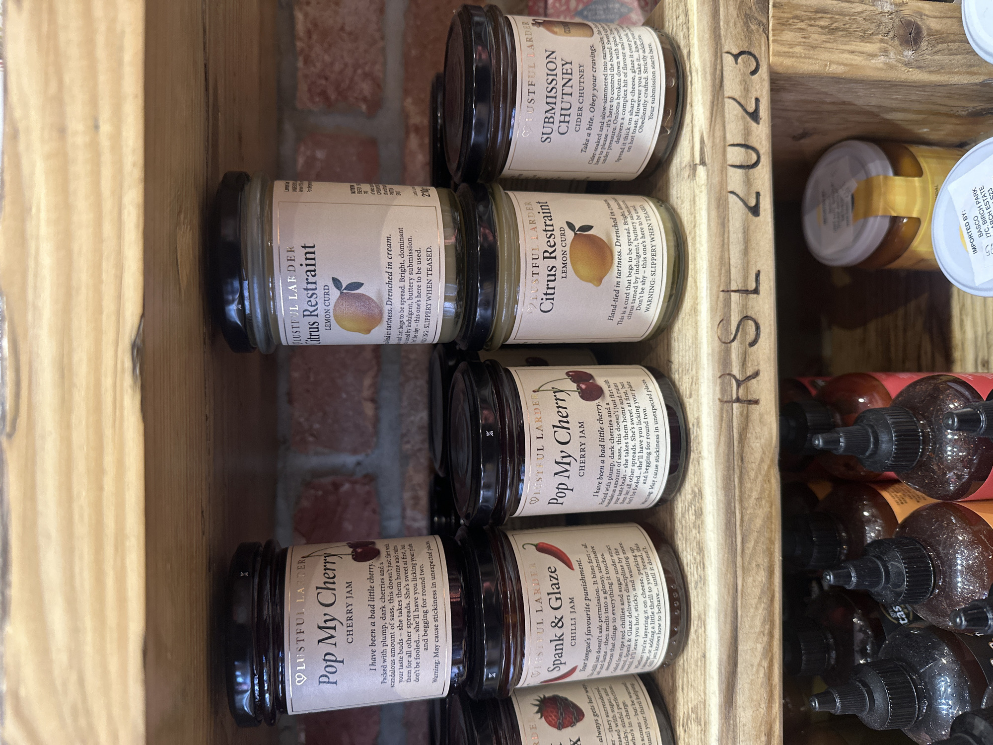 The image shows a wooden shelf in a shop, presumably in St Albans, United Kingdom, containing multiple jars of Lustful Larder artisanal condiments. There are visible jars of "Pop My Cherry" cherry jam, "Spank & Glaze" chilli jam, "Citrus Restraint" lemon curd, and "Submission Chutney" cider chutney. The jars are neatly arranged, showcasing the labels with descriptive text. A partially visible jar with a yellow label is also visible behind the Lusterful Larder products, and further back, a collection of bottles with black caps are also visible. The shelf itself is marked with "RSL 2023". A brick wall is visible in the background.