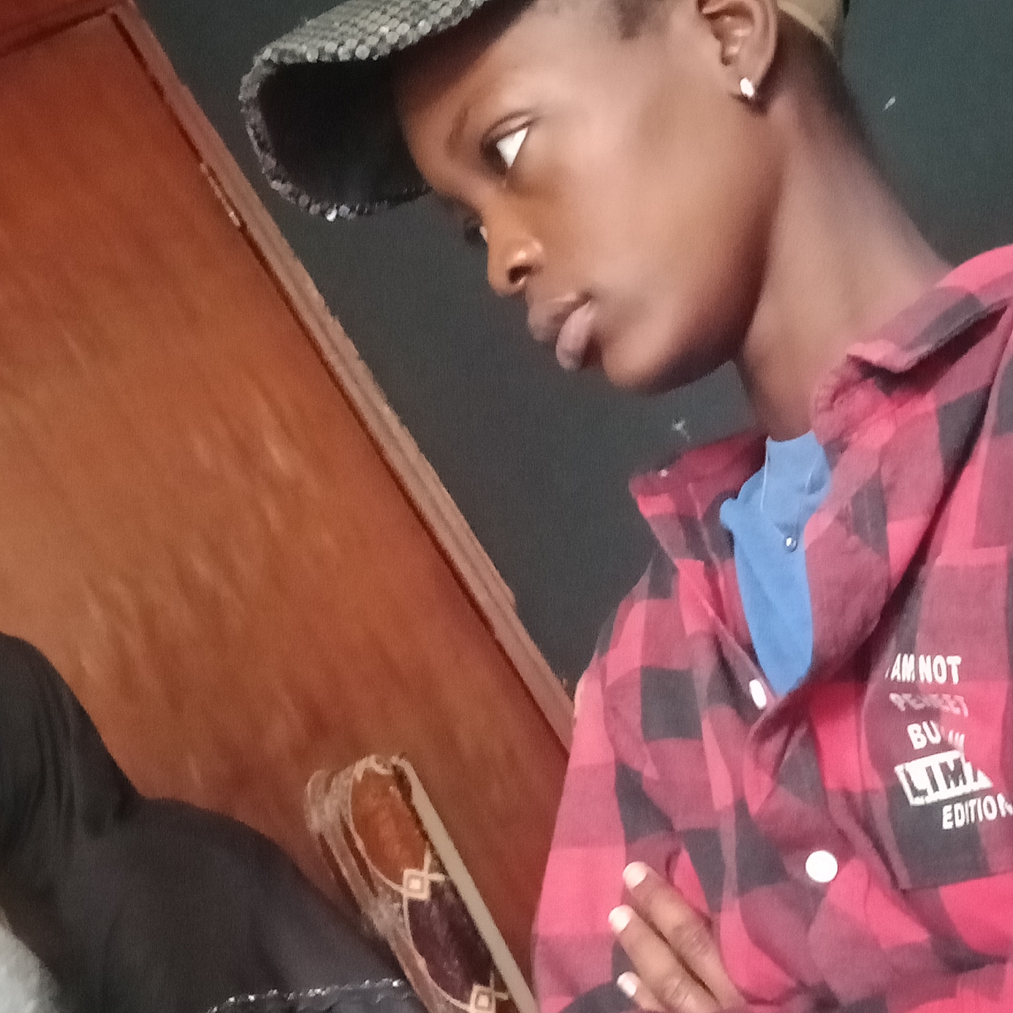 A young person wearing a red and black plaid shirt and a black patterned cap is captured in a close-up, profile shot. They are looking downwards to their left. A light blue undershirt is visible beneath the open collar of the plaid shirt. A silver stud earring is worn on the visible ear. The background is split between a dark surface and a wooden surface. A portion of a black garment is visible on the left side of the frame. Text is visible on the right side of the plaid shirt, reading "AM NOT", "BU", and "LIMA EDITION". This image was taken in Mowe, Nigeria.