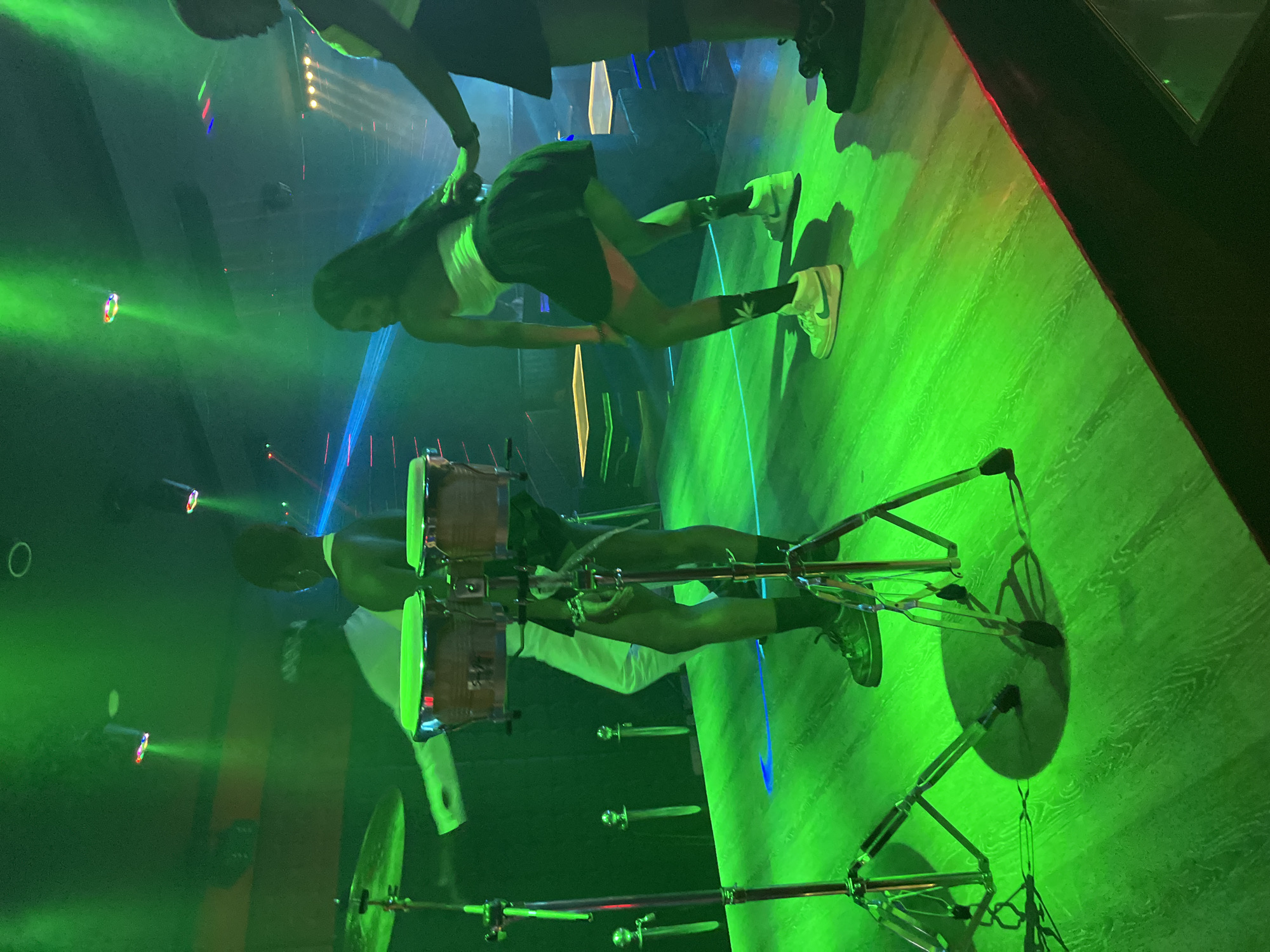 The image depicts a dimly lit nightclub or performance venue in Howayi, Nigeria, bathed in vibrant green stage lighting. In the foreground, a set of drum stands with cymbals and what appear to be bongo drums are visible. Three individuals are engaged in activity on the stage. To the left, a person is partially obscured, facing away from the camera and wearing white pants. In the center, a person with dark hair is dancing, wearing a white strapless top and a dark, short skirt, with white high-top sneakers. To the right, another person, also with dark hair, is visible from the back, wearing a dark skirt and dark shoes, with their hand extended. The stage floor is a glossy wooden surface reflecting the green lights. Several vertical, sword-like objects are placed along the back wall of the stage. The overall atmosphere suggests a live performance or entertainment event.