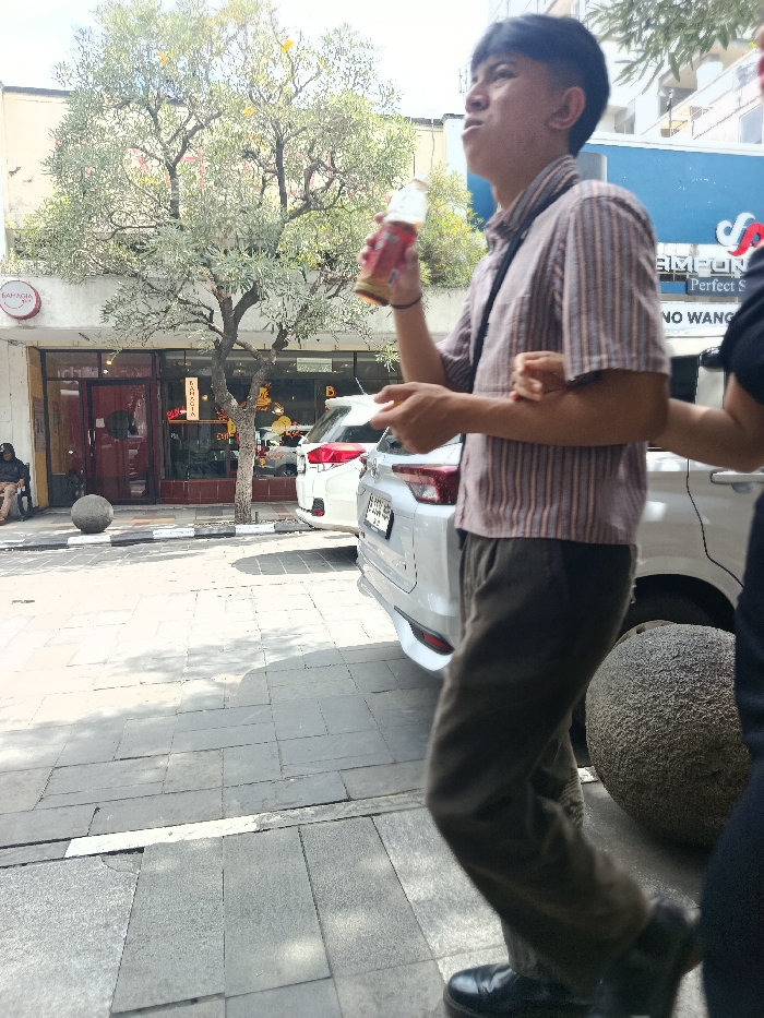 A young man, wearing a striped shirt, brown pants, and black shoes, is walking on a sidewalk in the city of Bandung, Indonesia, located at -6.918 latitude, 107.61 longitude. He is holding a brown glass bottle in his right hand and looking upward. Another person, whose face is not visible, is partially obscured by the camera's frame and is standing behind the man. The man is walking toward the right side of the image. In the background, there is a large tree, a parked white car, and a sign with the word "SAMPOEN" on it. The environment is a pedestrian area with gray brick sidewalks and a large, rough, gray rock on the right side of the image. The man is walking past the car, which is parked close to the sidewalk. There is a building with a storefront behind the car, and the front of the building has large windows and a brown door. A person sitting down is visible in the window, but the details are obscured. There is a large tree in front of the storefront. The image suggests a sunny day, as shadows are visible on the sidewalk. There is a small, round, gray stone next to the sidewalk, and a white line is painted on the edge of the sidewalk. The sidewalk is lined with gray brick pavers. The man is walking in the middle of the sidewalk, with the large rock on the right and the parked car on the left.