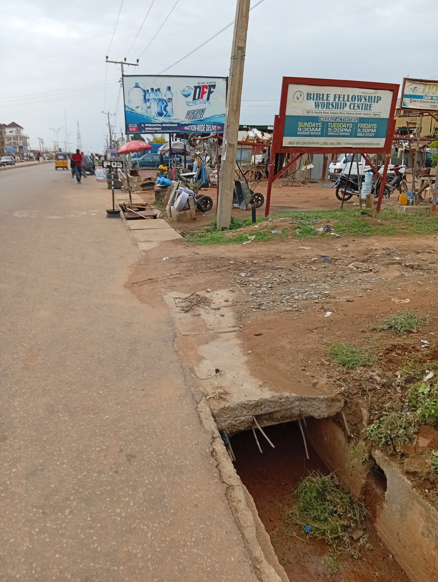 The image depicts a street scene in Sabon Tasha, Nigeria, near the Bible Fellowship Worship Centre at KM 10 Kachia Road. A damaged concrete drainage ditch is visible in the foreground, with exposed rebar and debris inside. A road runs alongside the ditch. Further down the street, a "POS" stand is set up near a bicycle repair area, and a man is walking. In the background, a large advertisement for DFF table water is visible.