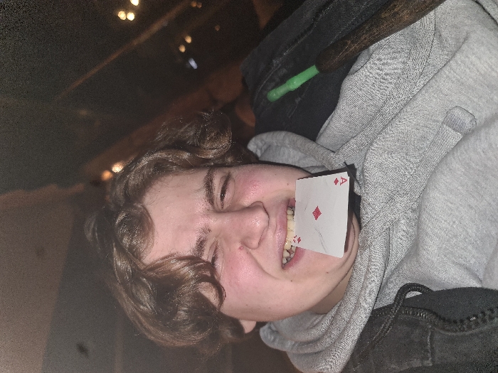 A person with curly brown hair and a fair complexion is shown with the Ace of Diamonds playing card stuck between their teeth. They are wearing a gray hoodie with a dark jacket partly visible in the frame. The lighting is dim, and the photo is slightly out of focus. The background is dark and blurry, with some faint light sources visible. The image appears to be taken from an unusual angle, possibly tilted.