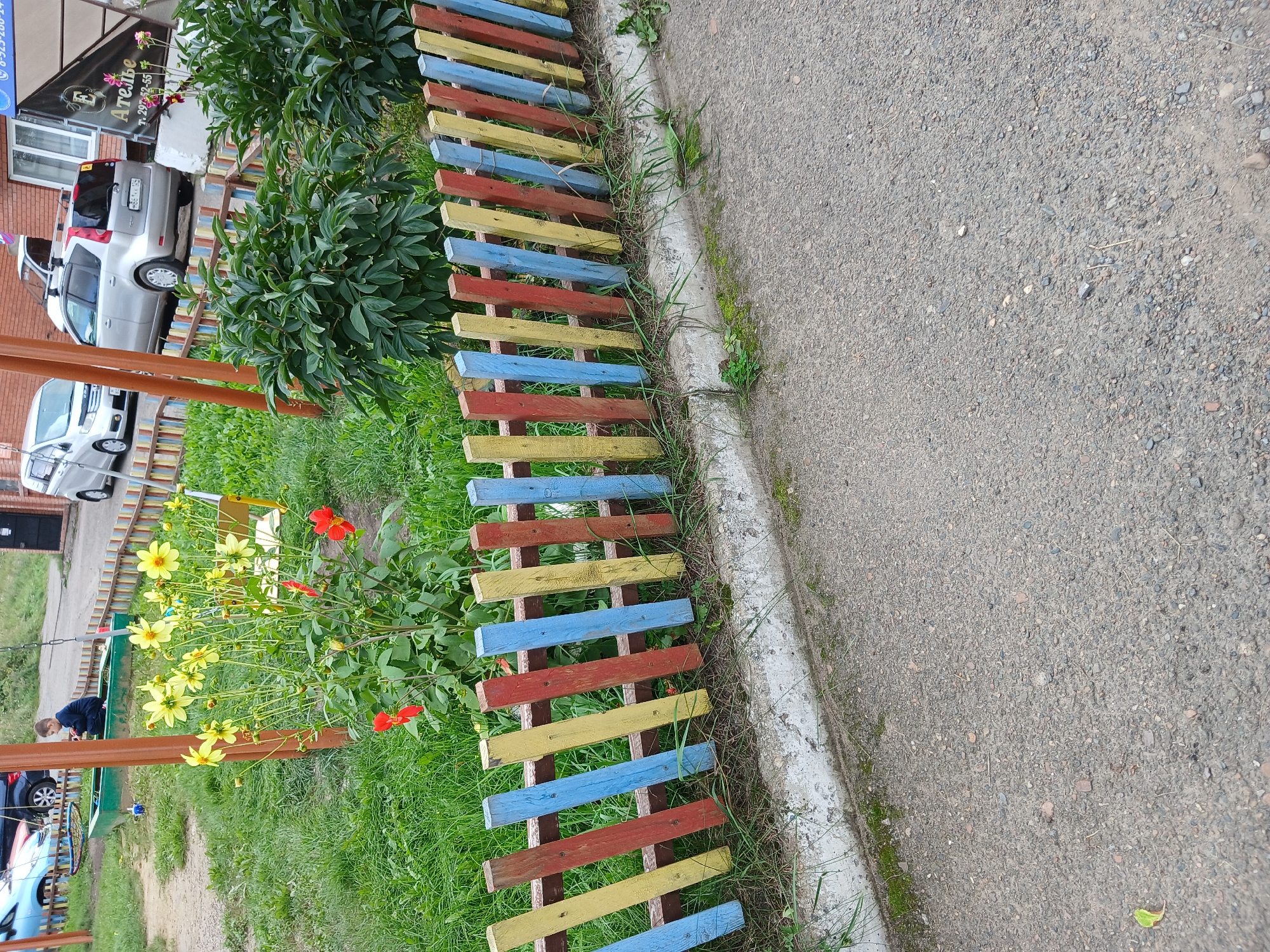 The image depicts an outdoor scene in an urban environment, possibly a playground or a residential courtyard. In the foreground, a colorful, low wooden fence made of vertical slats painted in red, yellow, and blue borders a patch of grass. Beyond the fence, yellow and red flowers are in bloom, with green bushes to the right. In the background, a young boy is sitting in a green sandbox. Several cars are parked in the vicinity, including a silver hatchback with a roof rack and a silver minivan. A building with windows is visible in the upper right corner, and signage with Russian text is also present. The ground is a mixture of asphalt and dirt.