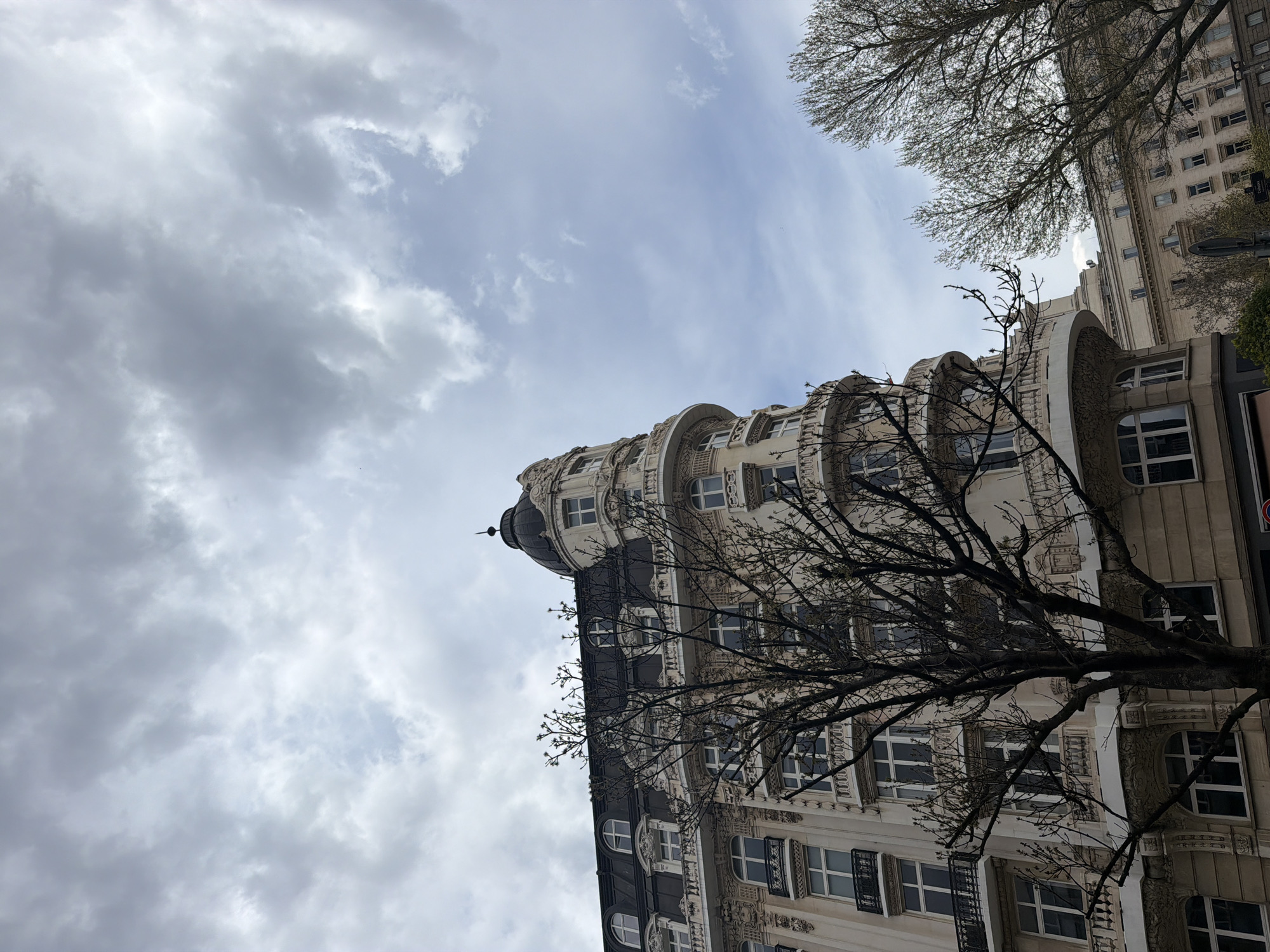 The image captures a grand, ornate building in Sofia, Bulgaria, from a low-angle perspective, looking upwards towards the sky. The primary subject is a multi-story building with elaborate architectural details, possibly in an Art Nouveau or Art Deco style. Its facade is light-colored, featuring numerous windows, some rectangular and some distinctive rounded bay windows with decorative carvings. Darker rooflines or sections of the building are visible, adorned with intricate ironwork on balconies. A domed turret with a pointed finial crowns one corner of the building, adding to its unique silhouette.
In the foreground and mid-ground, the bare, intricate branches of several deciduous trees extend across the frame, partially obscuring the building and reaching towards the sky. The trees show nascent buds, suggesting late winter or early spring.
The sky fills a substantial portion of the upper and left areas of the image. It is largely overcast, featuring a mix of heavy, textured grey clouds alongside brighter patches of a lighter blue-grey. Another tall, less ornate building is visible in the upper right background, partially framed by tree branches.
The scene depicts an urban setting during daytime, under a cloudy sky, contributing to a slightly moody or contemplative atmosphere. There are no people visible, and no discernible text appears in the image.