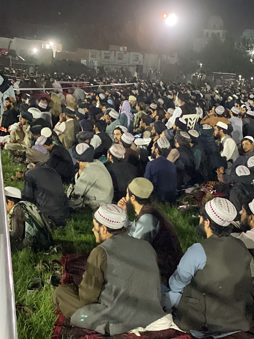 A large crowd of men sit outdoors at night in what appears to be Kandahar, Afghanistan. Many wear traditional pakol hats and some have beards. The scene is illuminated by bright lights, and a building can be seen in the background. The men are seated on grass and possibly rugs.