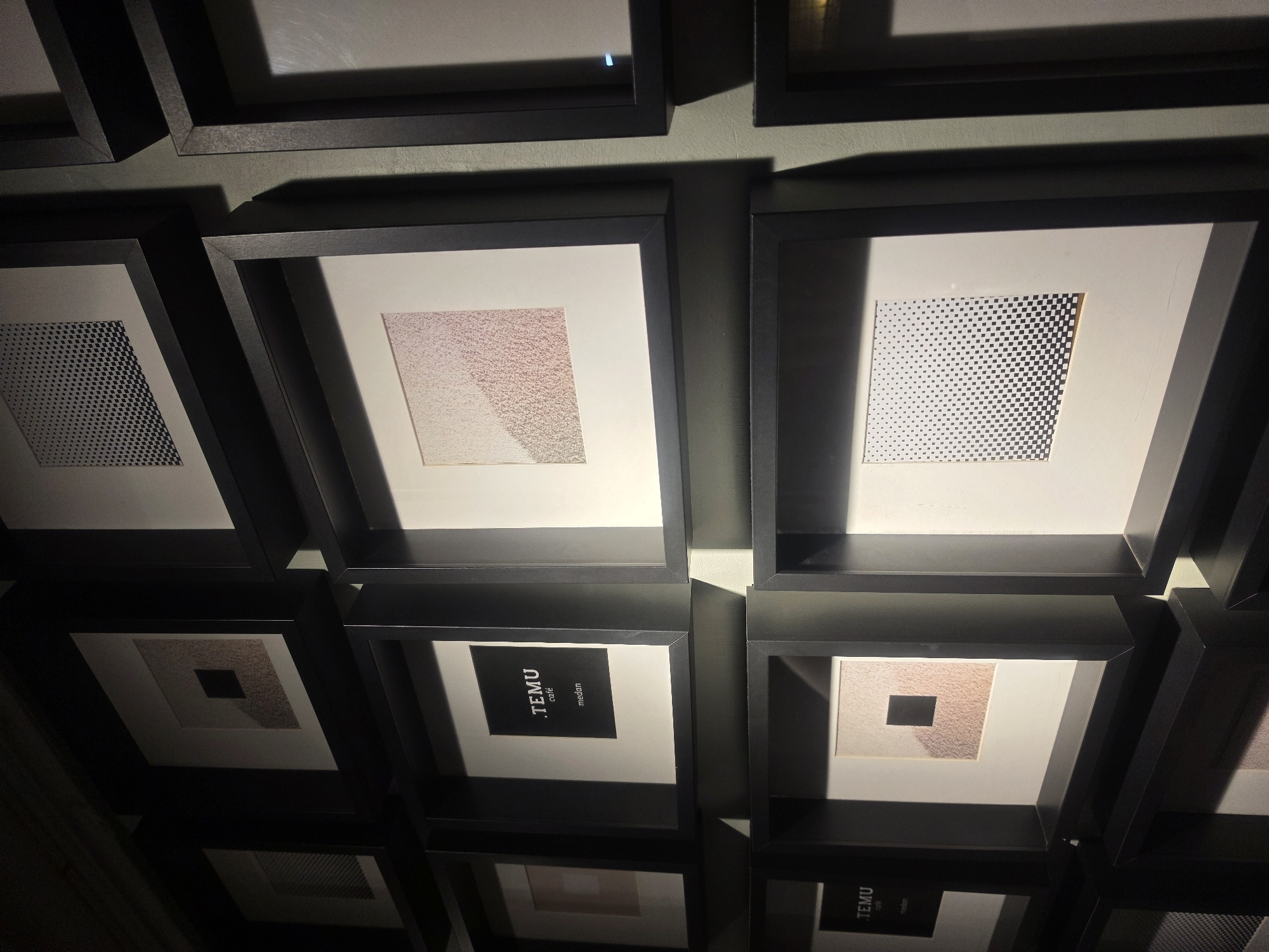 The image shows a wall with multiple framed artworks, likely a gallery or a cafe, in the city of Medan, Indonesia. The frames are dark, creating a contrast with the lighter matte borders and the artwork within. The artworks themselves are abstract and geometric, featuring patterns of dots, squares, and textured gradients. One frame contains text that reads ".TEMU café medan", indicating this is likely a cafe or restaurant named TEMU in Medan. The lighting in the scene is dramatic, with strong shadows cast across the artworks and the wall, suggesting directional spotlights or natural light entering from an angle. The overall impression is one of a modern and minimalist display of art.