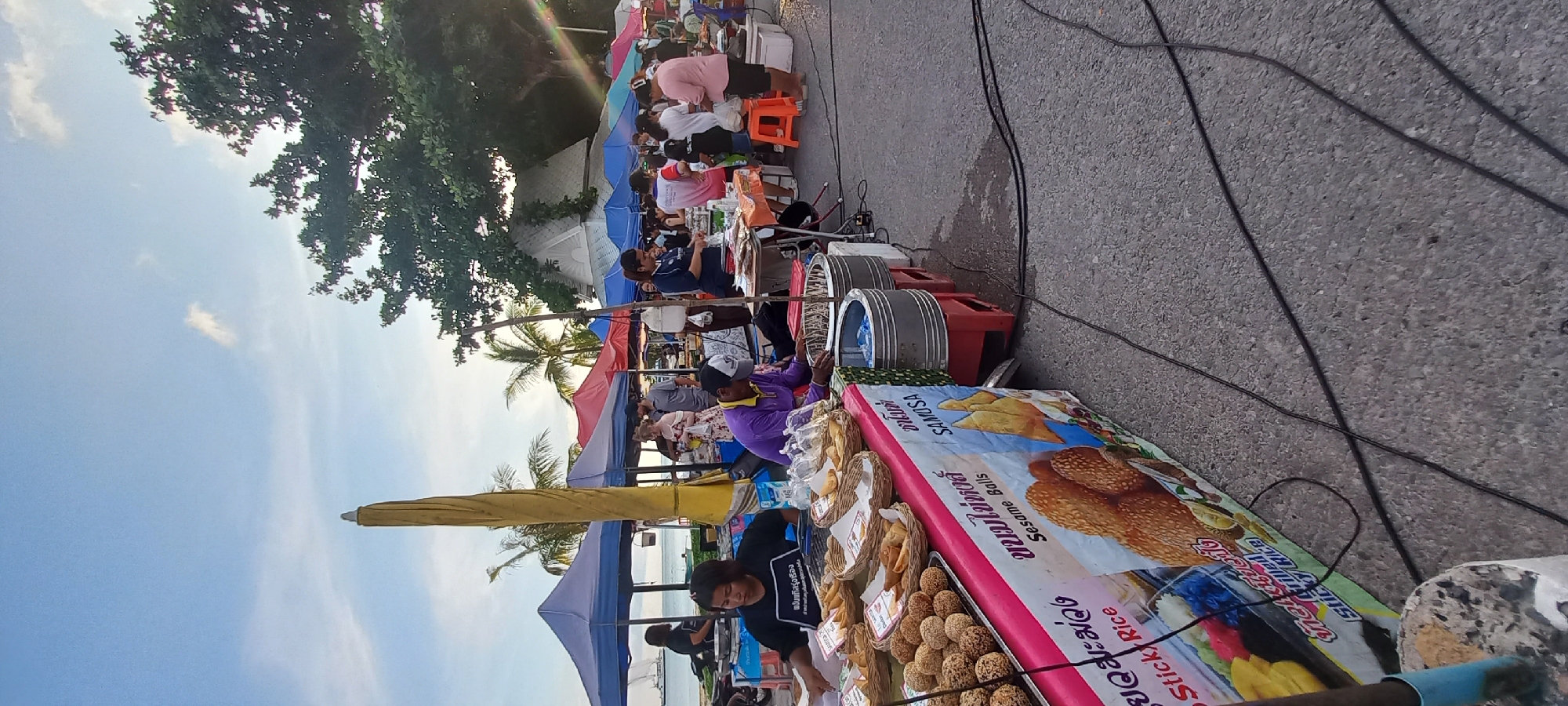 Here is a detailed description of the image:
**Scene:** A bustling street market scene, presumably in or near Thong Sala, Thailand, evident from the presence of Thai text on signage. The market is lined with vendors under colorful awnings, likely catering to tourists and locals.
**Environment:** The market appears to be located near a beach or waterfront, evidenced by a glimpse of water in the background, palm trees, and a clear sky. The surface of the street or the roadway appears to be asphalt.
**People:** There are several people present, including vendors and customers. The vendors are primarily focused on selling and interacting with customers. The customers are browsing and purchasing goods.
**Objects:** Market stalls are laden with items for sale, including what appears to be fried food items such as sesame balls and samosas, as well as other packaged goods. A variety of signage, some of which has Thai writing, are visible on the market stalls. Items such as plastic water bottles, umbrellas, and electric cables are also visible.
**Actions:** The vendors are actively selling, preparing, and handling food. Customers are examining items, making purchases, and engaging in conversation.
**Interactions:** Interactions between vendors and customers are prominent, indicating a commercial atmosphere.