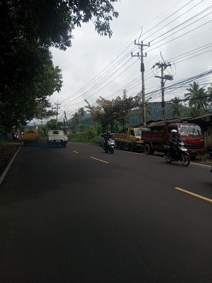 A rural road scene shows a yellow Toyota truck followed by a white Toyota pickup. A motorcycle rides in the same direction. Parked along the road are a yellow truck and a red dump truck. Another motorcycle is also on the road. Utility poles and wires cross overhead with trees and buildings along the roadside. Mountains are visible in the background under a cloudy sky.