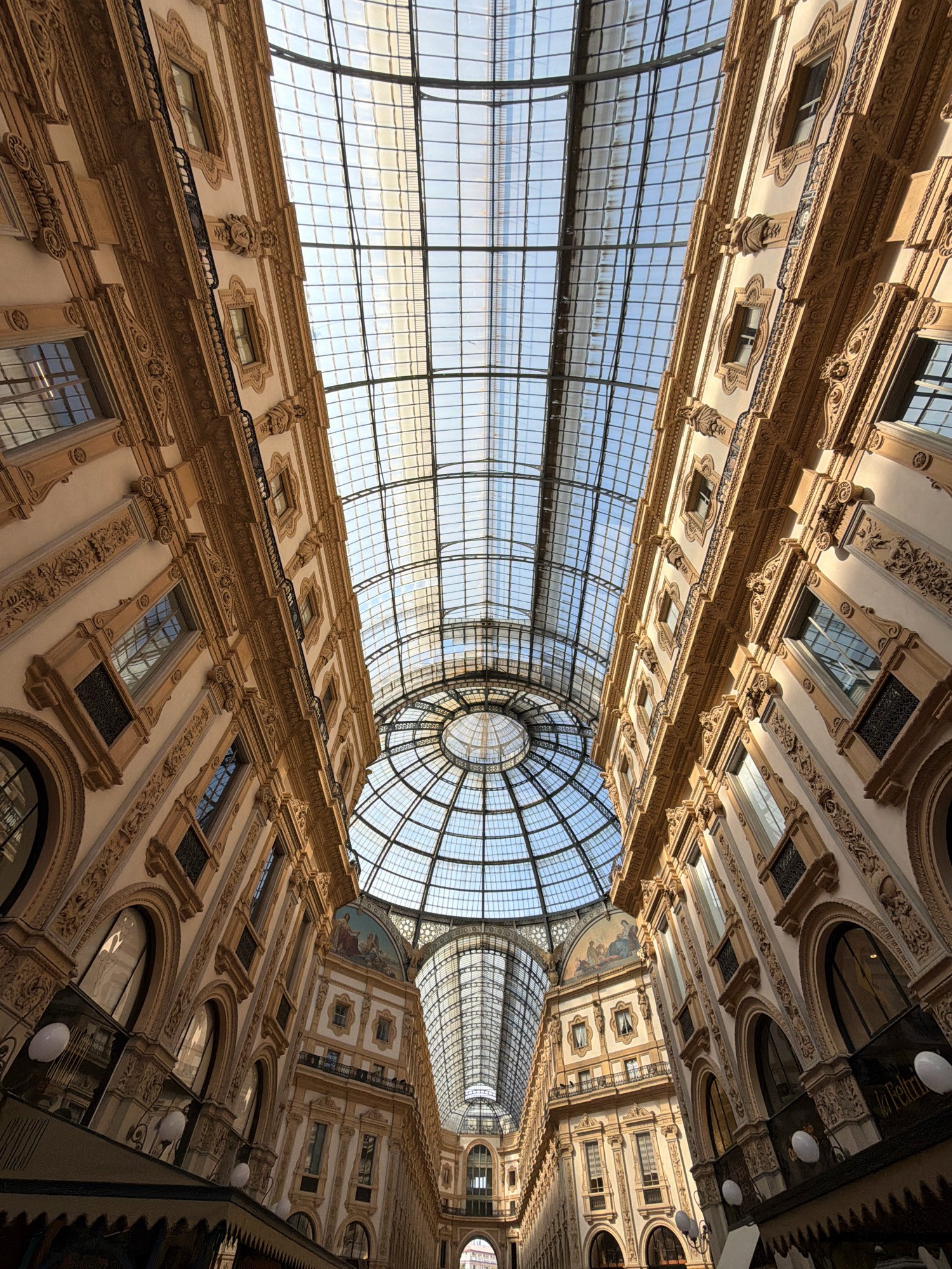 This media file shows an upward view of the Galleria Vittorio Emanuele II in Milan, Italy. The subjects are the grand architecture of the galleria, characterized by its ornate facades, arched windows, and elaborate decorative elements. The scene is an interior view of a large, covered shopping arcade with a magnificent glass and iron dome and a long vaulted glass roof that allows natural light to flood the space. There are no people clearly visible in the image.

The activity taking place is simply the display of the architectural magnificence of the galleria itself. It appears to be daytime, with bright sunlight filtering through the glass roof, creating a warm and inviting atmosphere. The sky visible through the glass is a clear blue.

Notable details include the intricate stonework and detailing on the building's exterior, the repetitive pattern of windows and balconies, and the impressive scale of the glass structures. The overall impression is one of grandeur, elegance, and historical significance.

There is no visible text in the image that is clearly legible. The location is identified as Milan, Italy, which is evident from the famous architecture of the Galleria Vittorio Emanuele II. The emotions evoked are likely awe and admiration for the beauty and craftsmanship of the building.