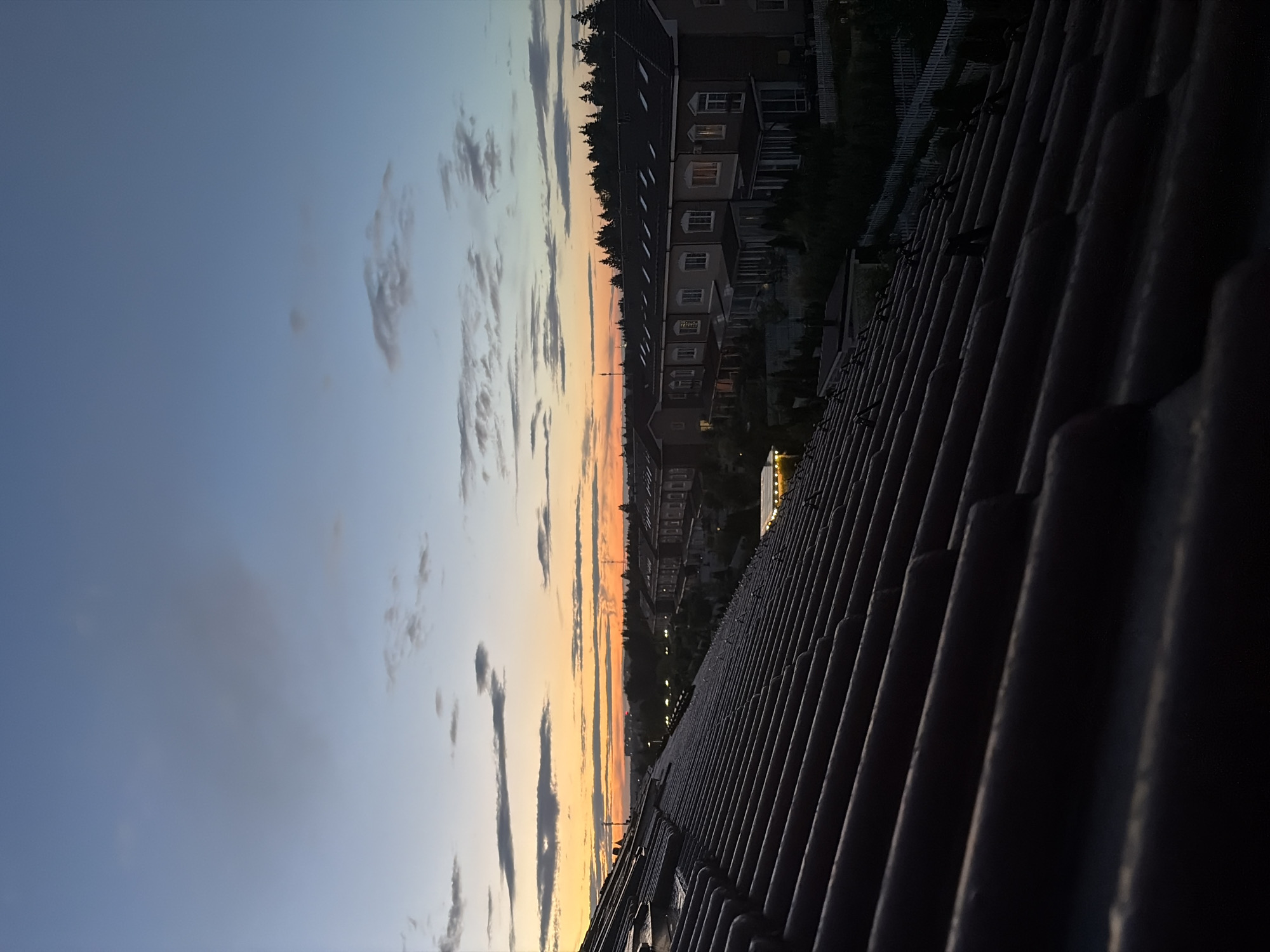 An elevated perspective captures a twilight scene over a residential area in Baranovo, Russia. The foreground is dominated by a dark, tiled roof, angled diagonally to the right, its texture and rows of tiles clearly defined in the dim light. Beyond the roof, a line of terraced houses stretches across the middle ground. These buildings are predominantly light-colored with dark roofs and evenly spaced windows, some of which are illuminated from within. A white picket fence runs along the front of the properties. Behind the houses, a dense line of dark green trees forms a backdrop, suggesting a wooded area. The sky above is a gradient of soft blues and grays, with streaks of orange and yellow near the horizon, indicating the setting sun. Scattered clouds, ranging from wispy to more substantial, drift across the sky. The overall impression is one of a quiet, suburban evening. There are no people or animals visible in the image.