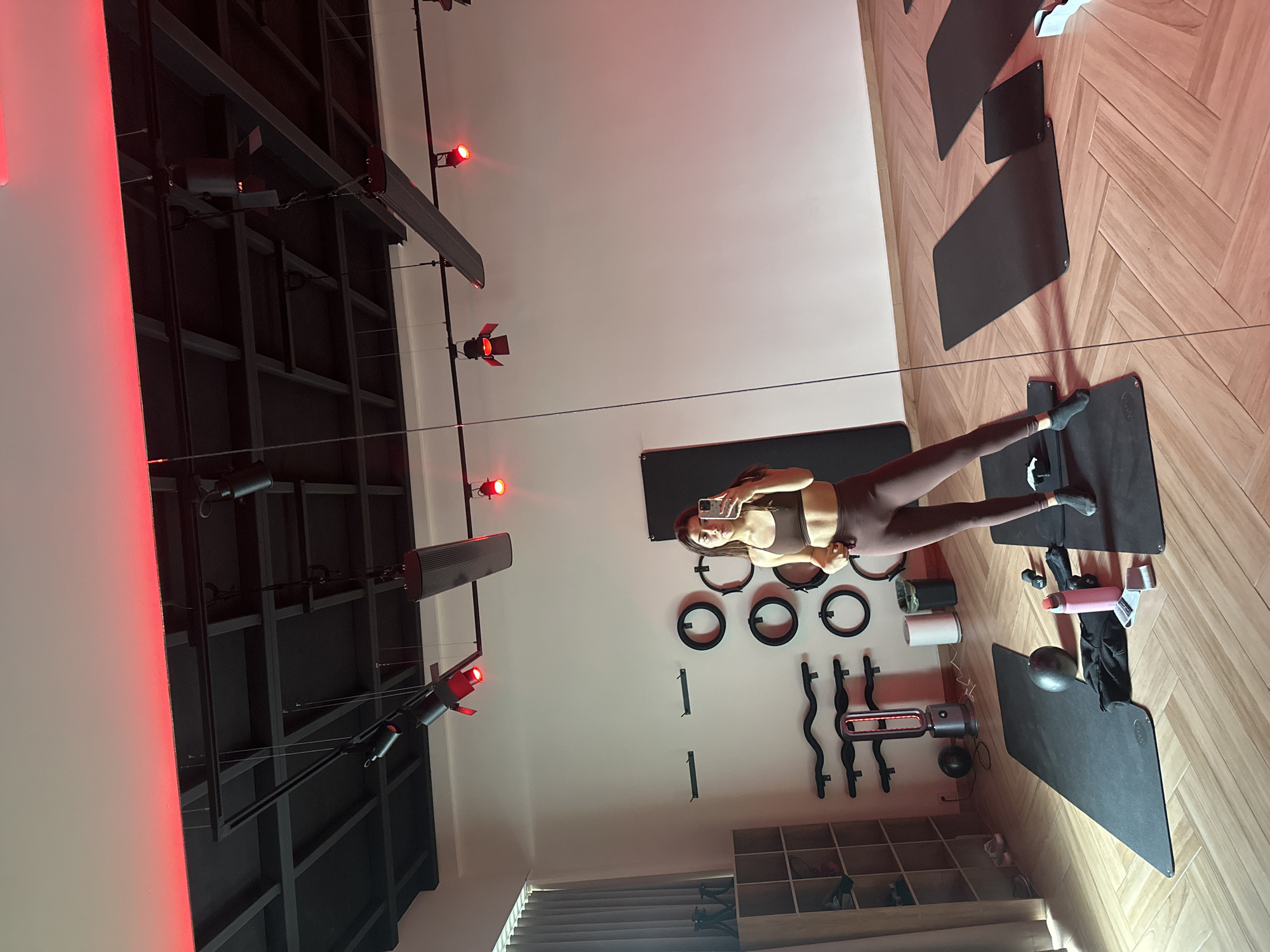 The media file shows a fitness studio with a woman in athletic wear taking a selfie. The studio features wooden flooring with a herringbone pattern, and several yoga mats are laid out. On the wall, there are various fitness accessories such as resistance rings, weights, and what appears to be a Pilates reformer. Red lights are strategically placed around the studio, casting a dramatic illumination. The woman is in a workout outfit, posing for the photo. The scene suggests an active or preparatory moment in a fitness session. The overall ambiance is modern and professional.