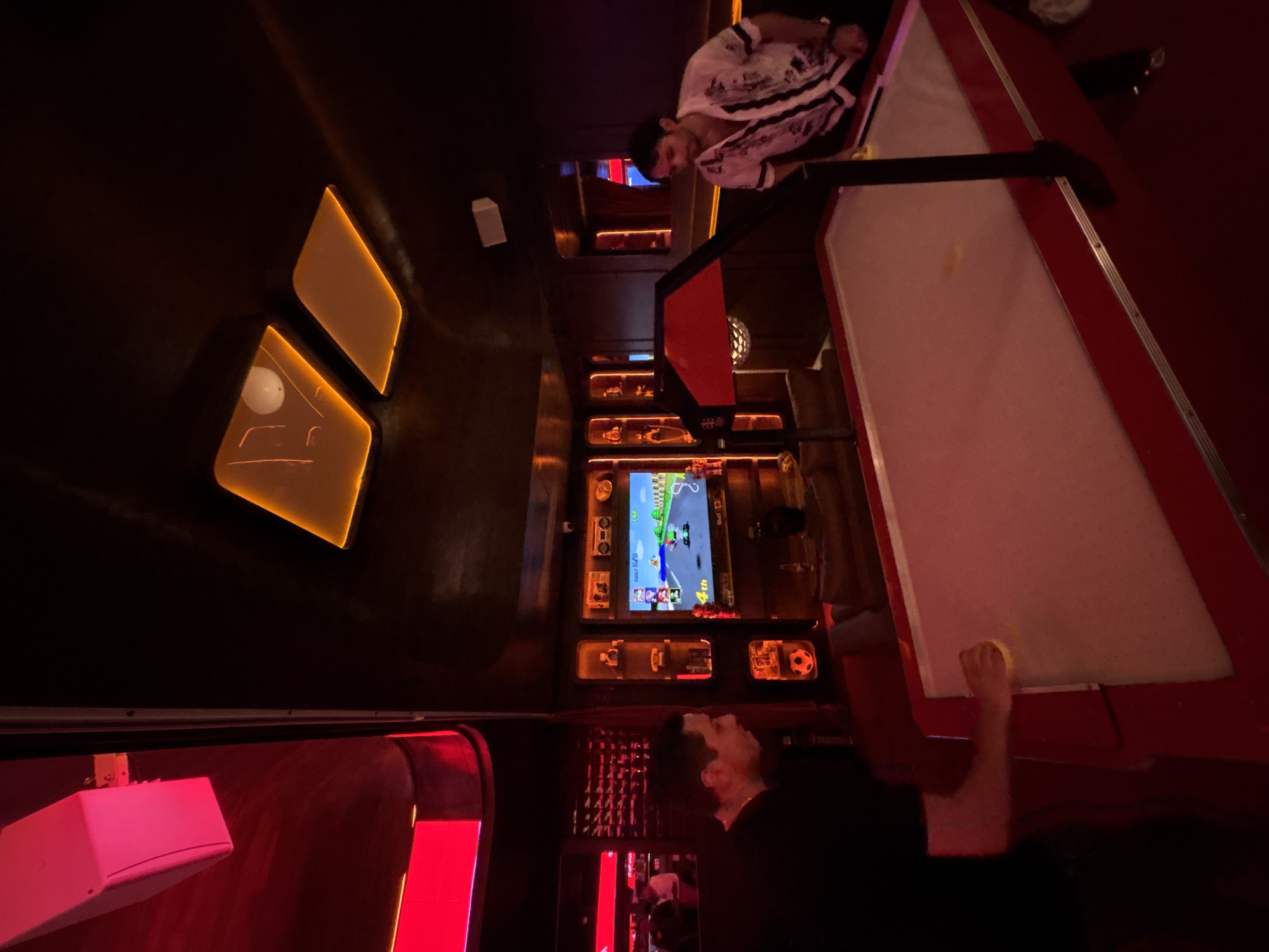 The image captures an indoor entertainment setting with dim, atmospheric lighting. Two men are engaged in gaming activities.
In the mid-ground, a man wearing a white shirt with a black pattern is leaning over a red and white air hockey table, seemingly focused on the game. His arm is extended towards the table. Directly below him, another man in a dark shirt is partially visible, reaching upwards, possibly also participating in the air hockey game or another activity.
The background features a wall adorned with illuminated shelves and a prominent video game screen. The screen displays a racing game, reminiscent of Mario Kart, showing a track with multiple racers and displaying "150" and "4th" (likely indicating speed and player position). Below this screen, various objects are displayed on shelves, illuminated by warm orange lights. These objects include what appear to be miniature vehicles, a small soccer ball, and other decorative items.
The venue is characterized by its dark walls and ceiling, punctuated by vibrant artificial lighting. Warm orange light emanates from rectangular fixtures on the left side of the image and from behind the shelves. Red neon or LED strips provide accent lighting, particularly visible on the bottom left and around the air hockey table. A white speaker or light fixture is mounted on the wall to the left of the man in the white shirt. A small disco ball is also faintly visible in the upper background. The overall atmosphere suggests a lively, dedicated gaming or recreational space, possibly an arcade or a bar.