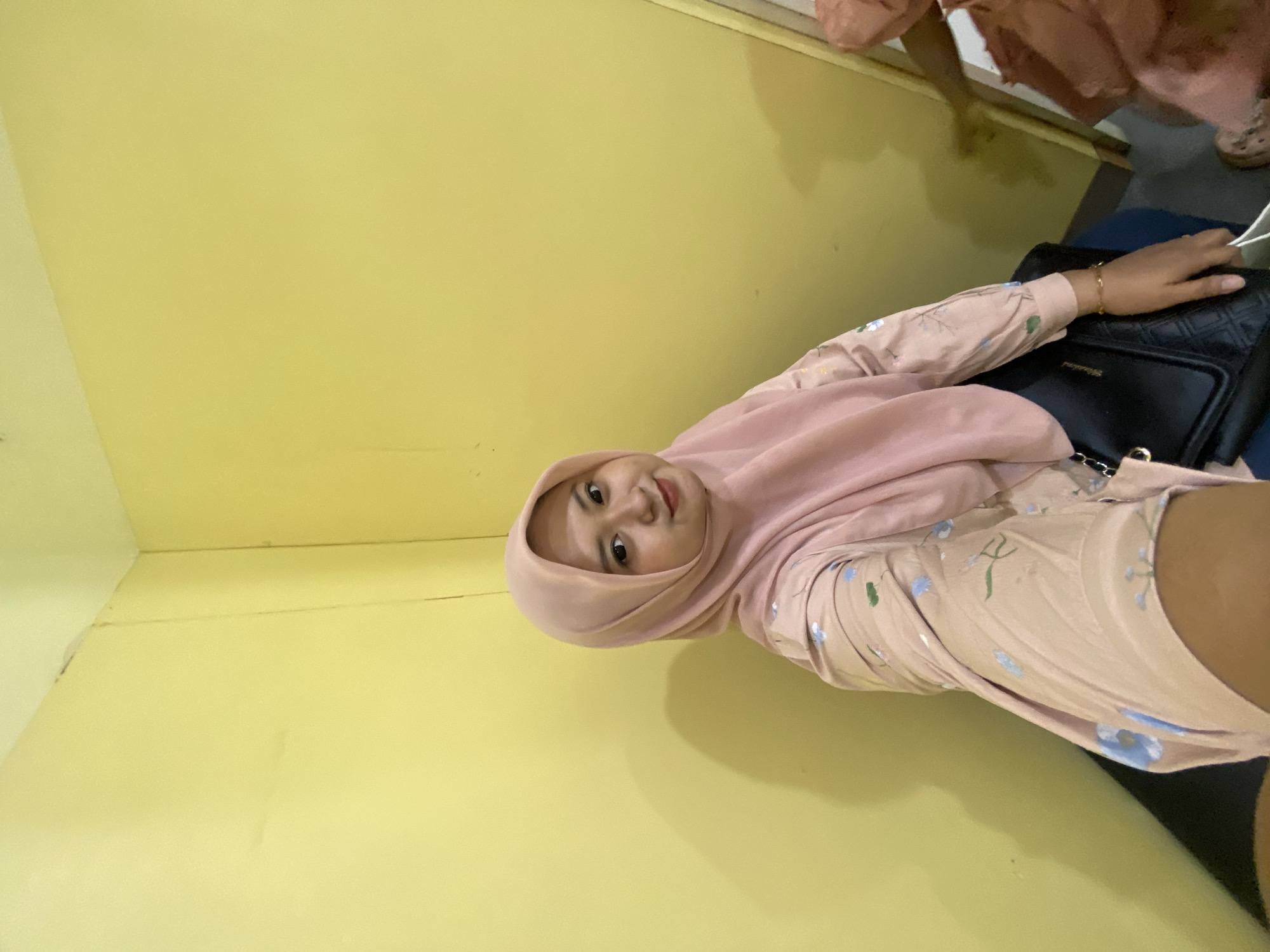 In Sidoarjo, Indonesia, a young woman wearing a pink hijab and a floral-patterned light pink blouse is captured in a selfie. She is seated, with her right arm extended, holding a white bag. Her left arm rests on a black handbag, which is placed on her lap. The background consists of a plain yellow wall. The lighting suggests an indoor setting, possibly a room. The image focuses on the woman and her immediate surroundings.