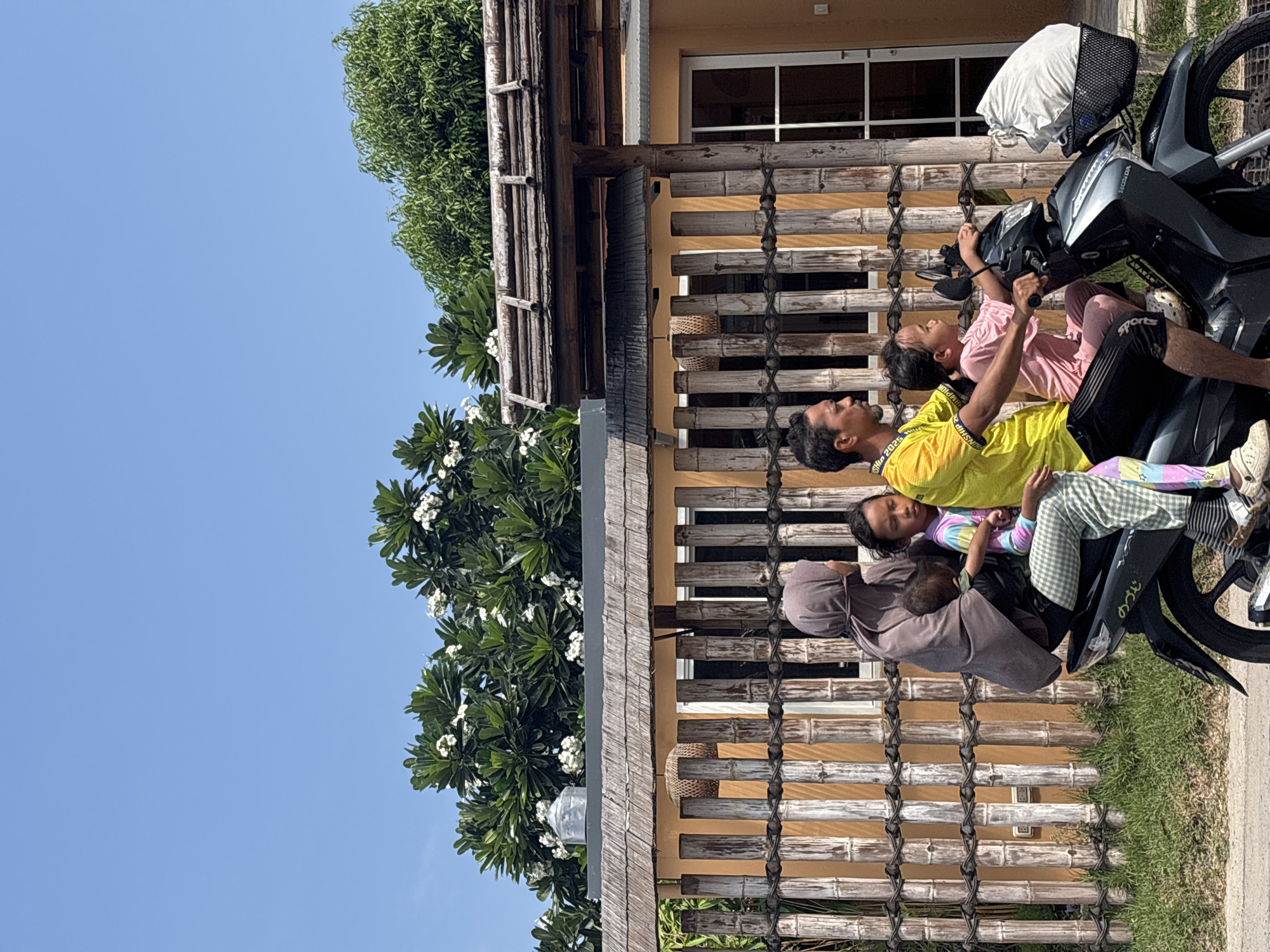 A man and three children are on a motorcycle in Choeng Thale, Thailand. The man, wearing a yellow shirt and black shorts, is in the middle. A child in pink pajamas is in front of him, on the motorcycle's handlebars. Behind the man is a child wearing a colorful top and checkered pants. The third child, dressed in a brown outfit, is at the very back of the motorcycle. A bamboo fence and a building with a window are behind them. Trees and a clear blue sky are also visible in the background.