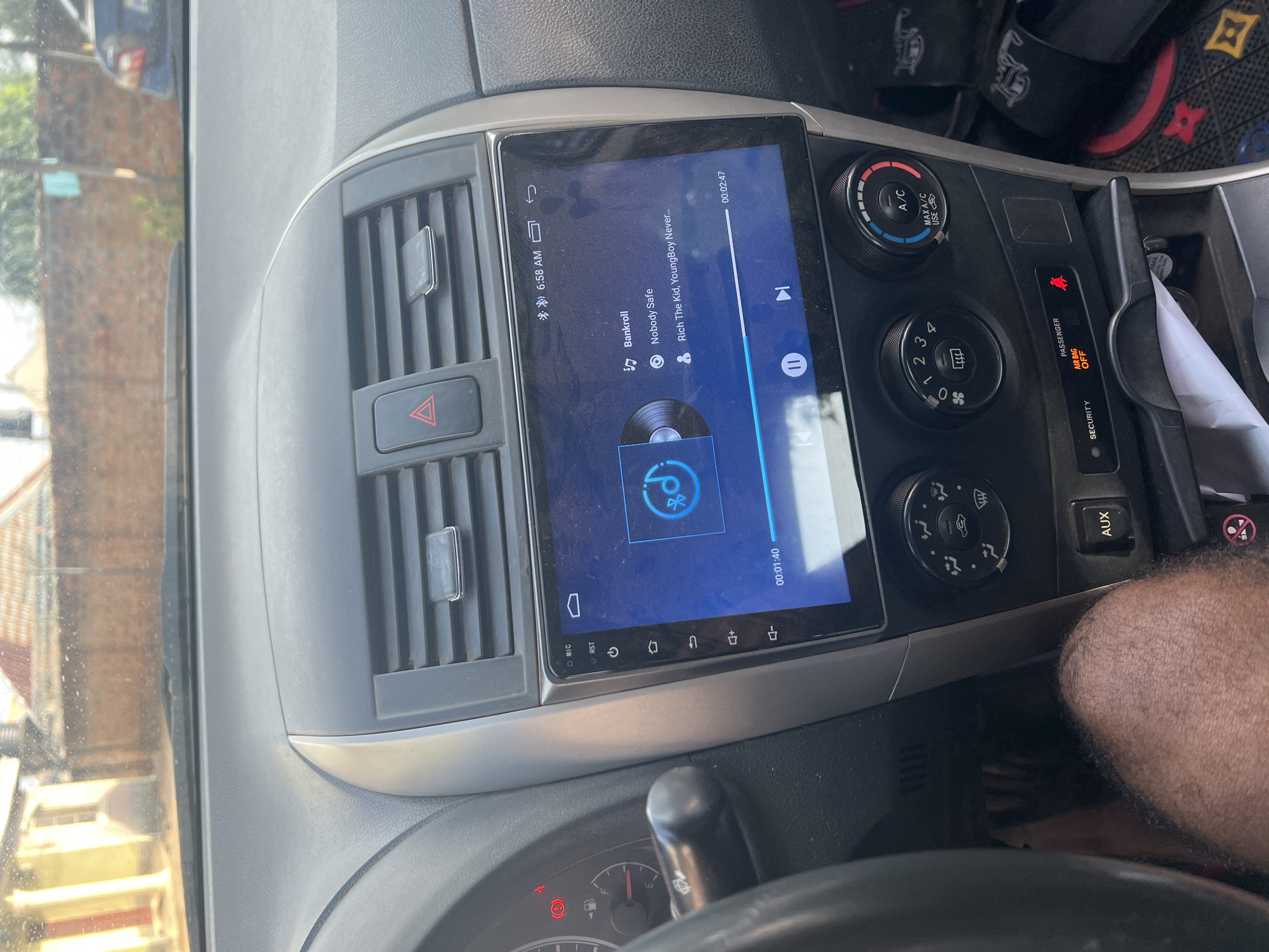 This image shows the dashboard of a car, specifically the center console area. The car is playing a song called "Nobody Safe" by Rich The Kid and YoungBoy Never Broke Again from the album "Bankroll" at 6:58 AM. The car's air conditioning and heating controls are visible, and the passenger airbag indicator is set to "off". There's a partial view of a person's hairy legs in the bottom right corner, along with a glimpse of the car's steering wheel. Based on the left side of the image, the car may be located in a city with greenery, possibly Markurdi, Nigeria.