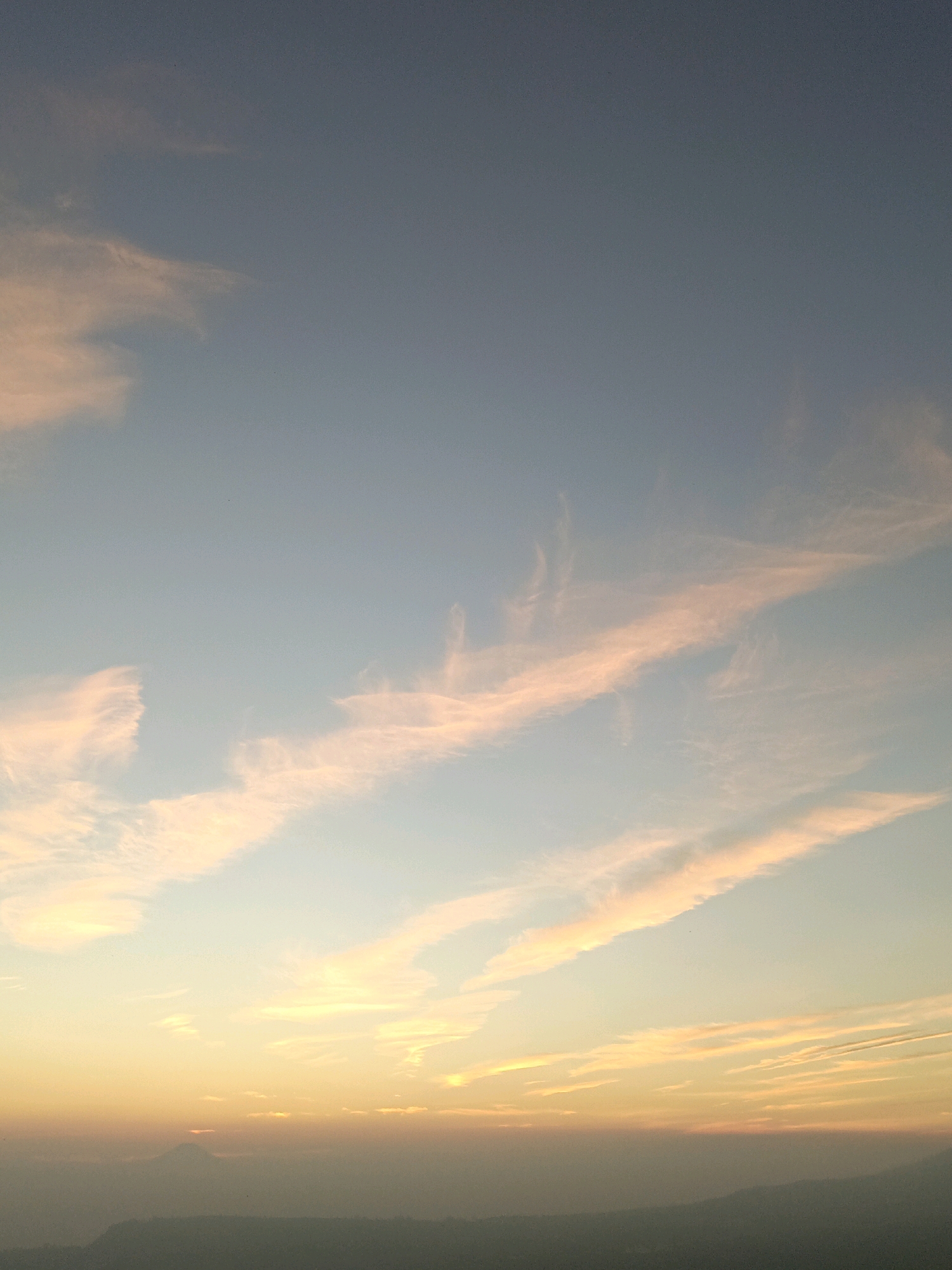 The image depicts a panoramic view of a sunset over the city of Butuh Kidul, Indonesia. The sky is filled with wispy clouds tinged with hues of pink and orange, transitioning into a soft blue as the eye moves upwards. Below the sky, a hazy landscape is visible, hinting at mountains or hills. In the distance, a silhouette of a mountain peak can be seen, with a sliver of the sun just peeking over its summit. The foreground consists of a darkened, indistinct silhouette of the land, suggesting the city below is obscured by the fading light and atmospheric conditions. There are no people or explicit actions depicted in the scene. The overall atmosphere is serene and tranquil, characteristic of a sunset.