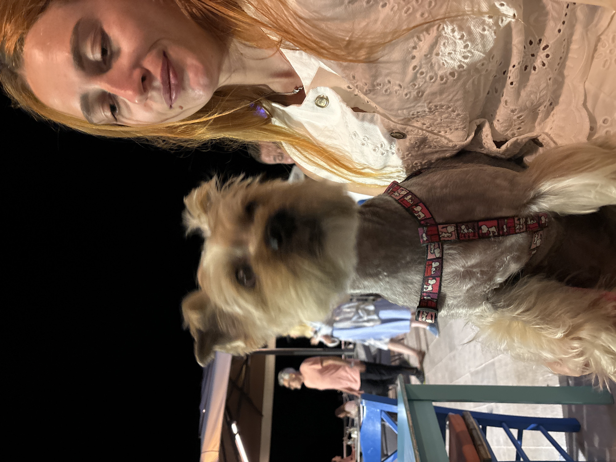 A medium shot captures a woman with long, reddish-blonde hair holding a small, light-brown Yorkshire terrier. The woman is wearing a white eyelet blouse and a silver necklace. The dog, wearing a red harness with a Snoopy print, is in the foreground and slightly out of focus. In the background, on a wooden deck, people are seen at tables under a large canopy. To the left, a blue table and chair are visible. The scene appears to be an outdoor dining area at night in Petrovac centar, Montenegro.