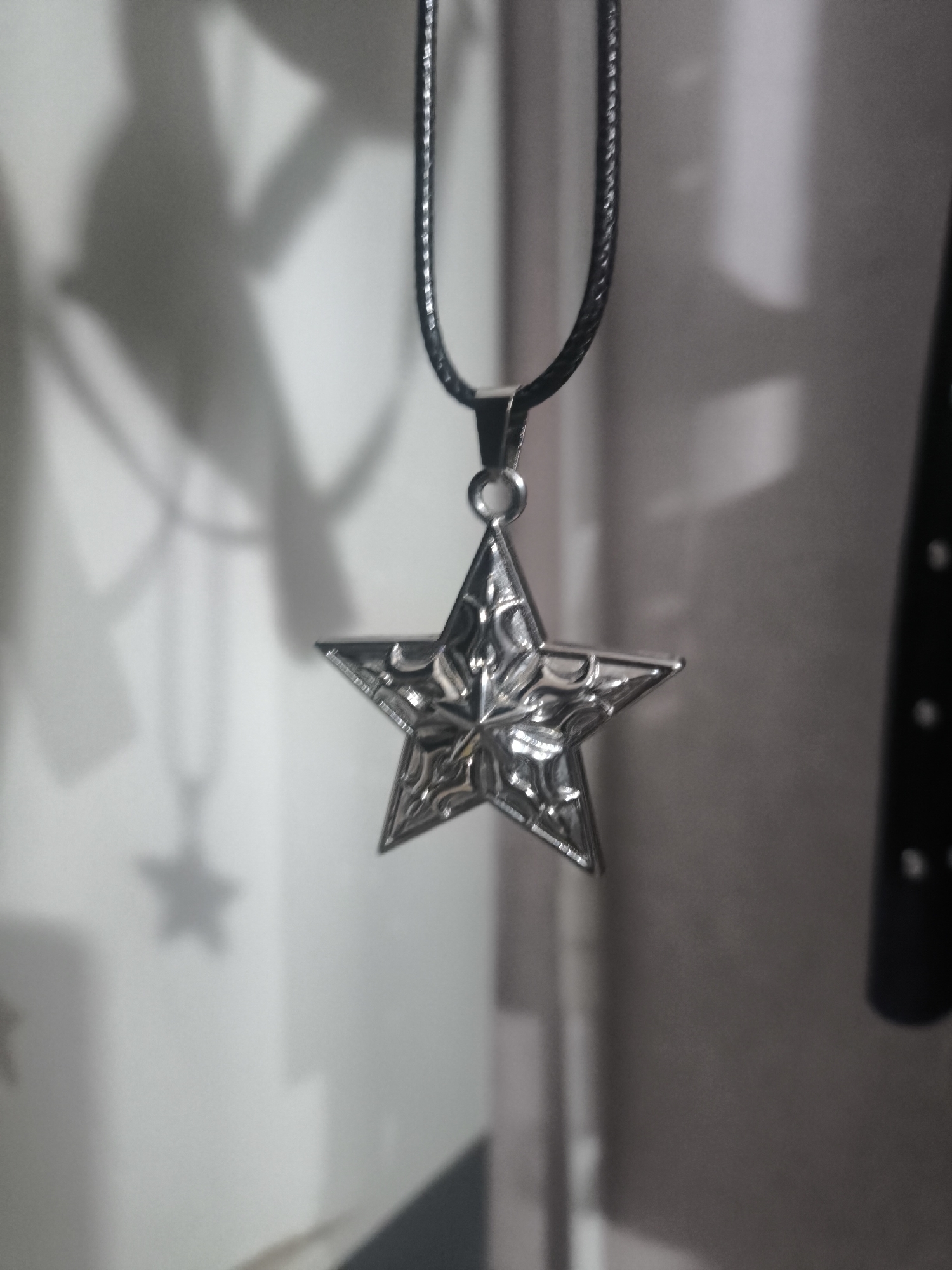 A star-shaped pendant with intricate detailing hangs from a black cord. The pendant is silver and appears to be made of metal. The background is out of focus, with soft shadows suggesting an indoor environment. There is no information about the location in Gentan, Indonesia.