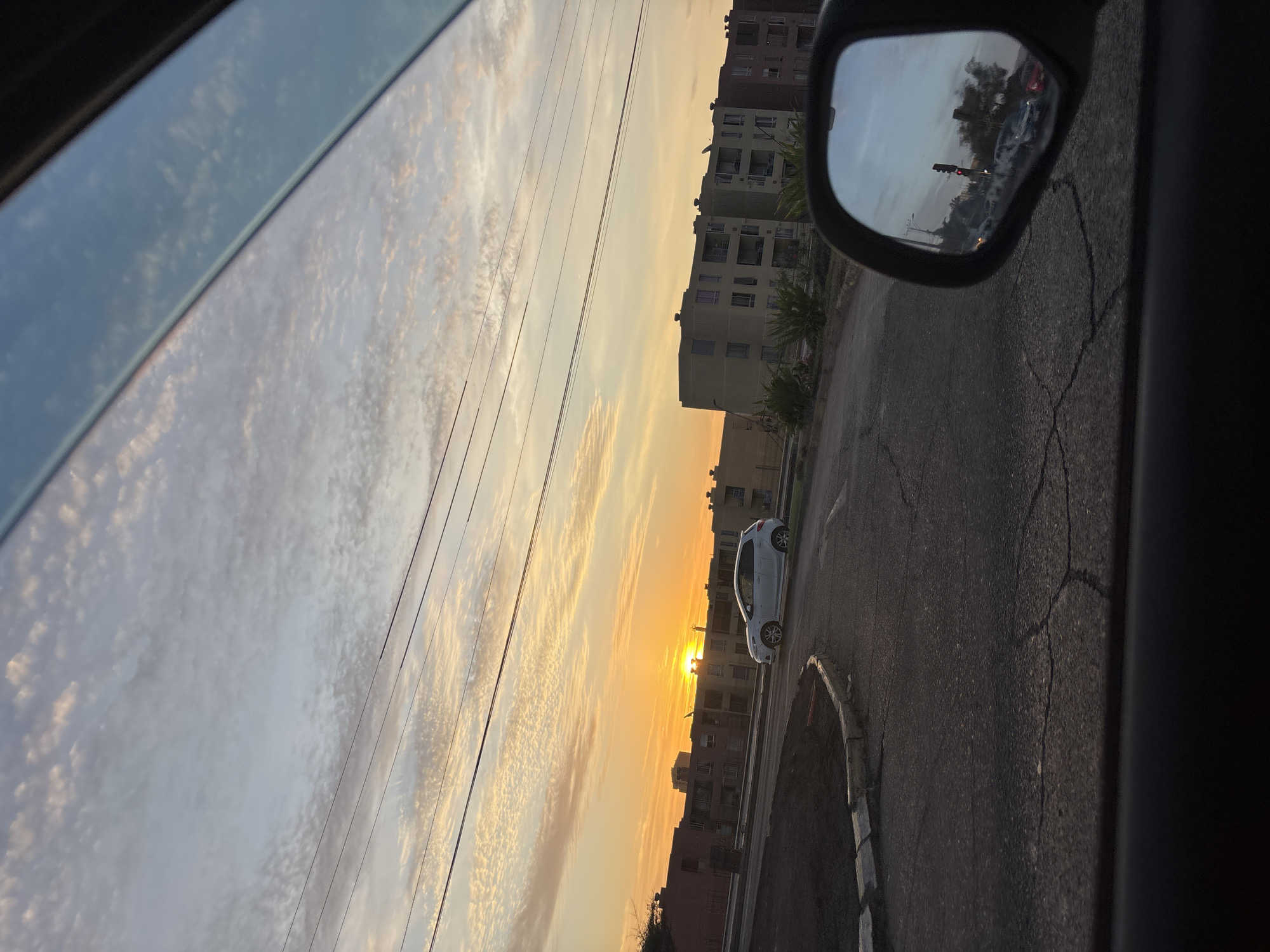An urban street scene in Arica, Chile, is captured from the interior of a vehicle, looking outwards through the side window at sunset. The vibrant sky dominates the upper left, displaying warm hues of orange and yellow as the sun dips towards the horizon. Scattered altocumulus clouds glow in the golden light, creating a textured pattern across the sky. Several power lines stretch across the frame.
Along the right side, the dark frame of the car's interior is visible, with a side-view mirror reflecting a glimpse of other vehicles and possibly a traffic light. Below the sun, a white car is parked on the cracked asphalt road. In the background, a row of multi-story residential buildings with multiple windows and balconies lines the street, silhouetted against the bright sky. The road surface shows signs of wear with visible cracks. No visible text is present.