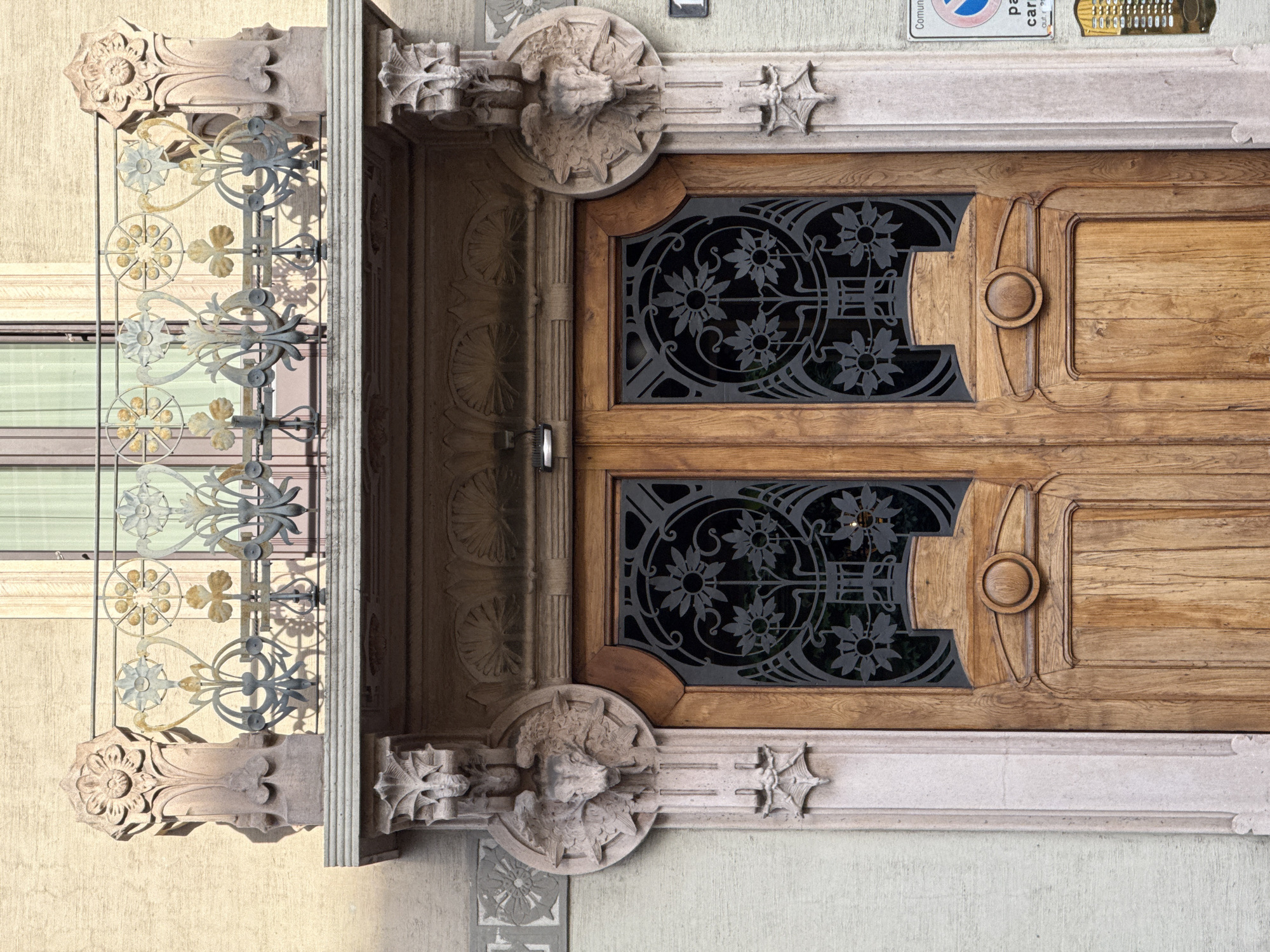The image captures an ornate, vintage entryway in Milan, Italy. The focus is on a pair of double wooden doors, richly decorated with black floral metalwork inserts in the upper portions. Each door features a round wooden handle. The doors are framed by an elaborate stone structure with intricate carvings, including floral motifs and sculpted figures. To the left of the doorway, an ironwork railing with a floral design is visible, partially obscuring the view of an adjacent window. Above the doorway, signage and other elements are present. The overall style suggests an architectural period from the late 19th or early 20th century.