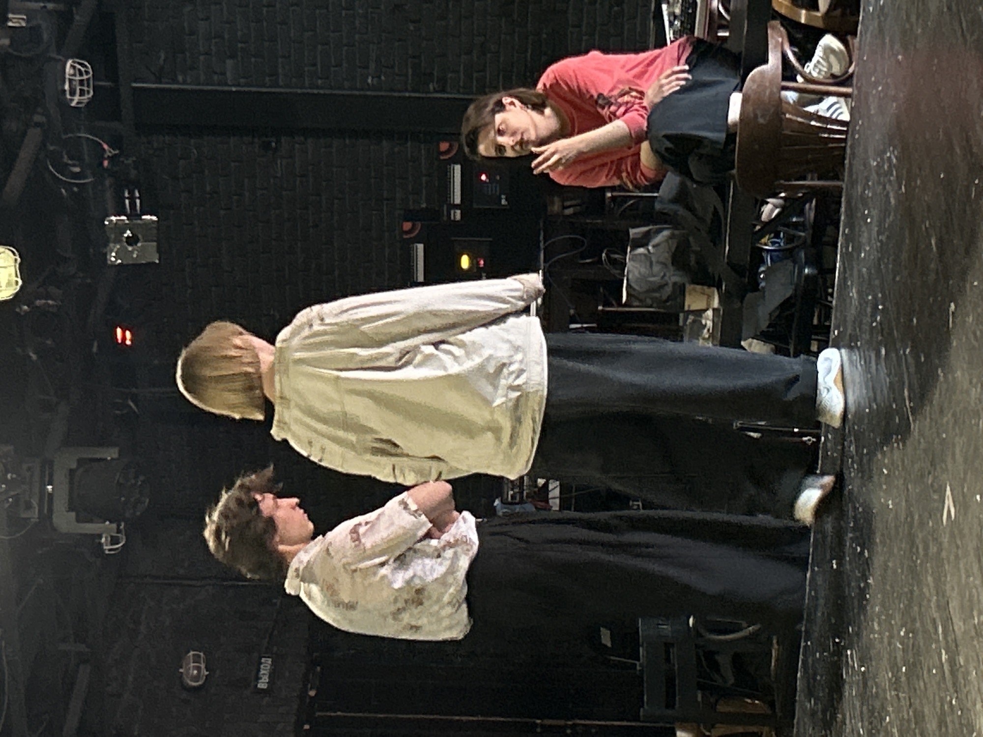 In a dimly lit indoor space with a black brick wall in the background, three individuals are present. Two people, appearing to be young adults, stand center stage. The person on the left is wearing a patterned short-sleeved shirt and dark pants, with their arms crossed. The person on the right is wearing a light-colored jacket and dark pants, with their back to the camera. A third individual, a woman, is seated on a chair to the right, wearing a coral-colored shirt and dark pants, gesturing with her hand. The environment includes stage lighting equipment overhead and various objects on a table in the background, including what appear to be electronic devices and bags. The floor is dark and scattered with debris.