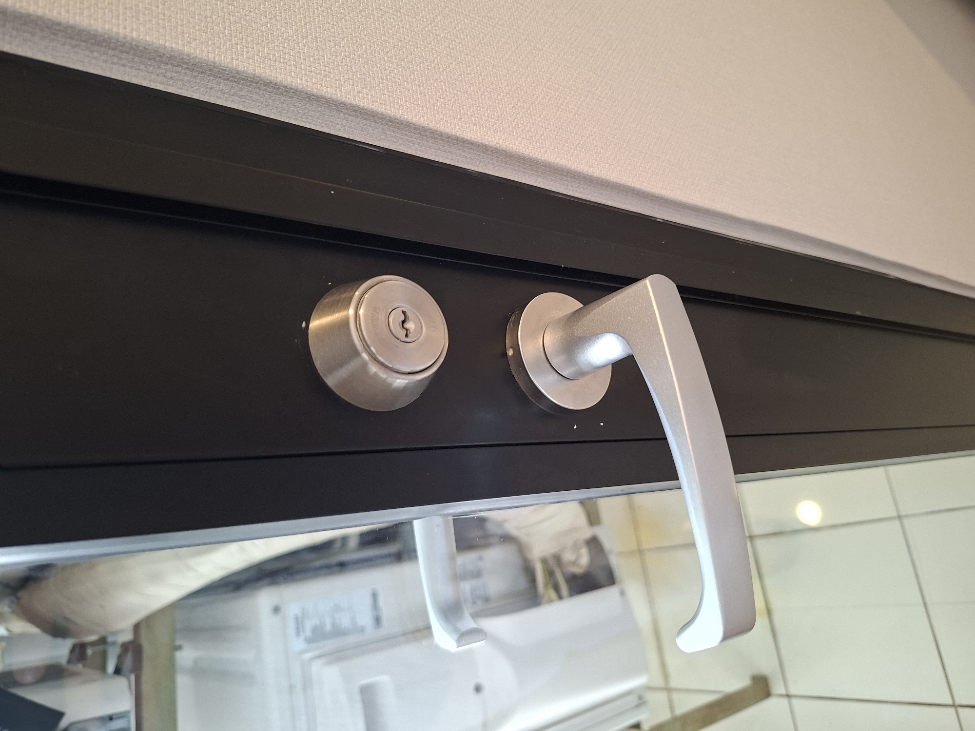 The image shows a close-up of a modern door lock and handle assembly. The lock is a deadbolt with a keyhole, and the handle is a lever-style mechanism, both made of brushed metal. The door itself appears to be dark gray or black, with a glass panel on the left side. Reflected in the glass are parts of what look like household appliances, possibly a washing machine, and tiled flooring. The wall to the right of the door has a textured, light-colored finish. There are no people visible in the image. The scene is likely an interior shot of a room or hallway in a residential or commercial building. It is impossible to determine the specific location in West Jakarta, Indonesia based solely on this image.