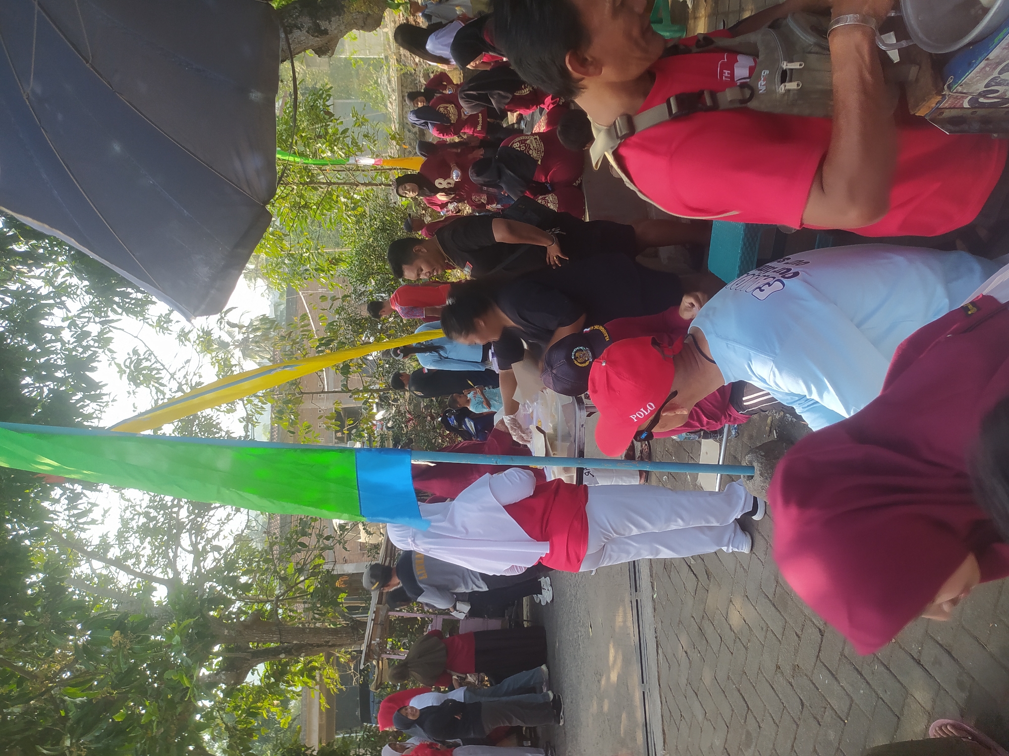The image depicts an outdoor gathering in Lopait, Indonesia, possibly a community event or festival. In the foreground, a person wearing a red shirt and a backpack is visible from the chest up, preparing something at a stall with a metal pot. To the left, another person in a light blue shirt and a red cap is seen from the back. Further left, a woman in a burgundy hijab is looking down. In the mid-ground, a group of people, some in red shirts and others in white attire, are gathered around a table. Several people are wearing hijabs. Tall flags, one green and blue, and another yellow, stand to the left of the frame. In the background, trees with lush green foliage line the area, suggesting a sunny day with dappled sunlight filtering through the leaves. A dark umbrella is visible over a stall on the right side of the image. The ground appears to be paved with interlocking bricks. The overall scene suggests a lively public event taking place in a semi-urban or village setting in Lopait.