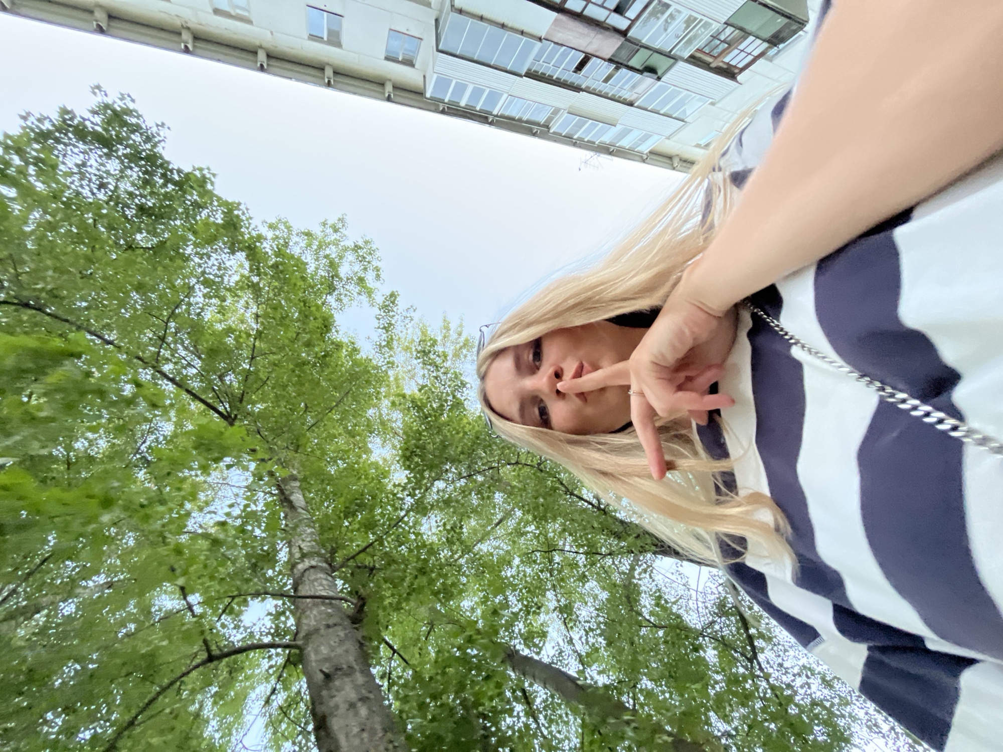 A low-angle shot captures a woman standing near a tall birch tree in the city of Chaikovsky, Russia. The woman, positioned in the upper right of the frame, is blonde and wears a striped navy and white shirt. She holds a finger to her lips as if gesturing for silence. Above and slightly behind the trees, the corner of a light-colored building with windows is visible. The sky is overcast. The trunk of the birch tree is visible on the left side of the frame, reaching towards the top of the image, with dense green leaves filling the upper left quadrant.
