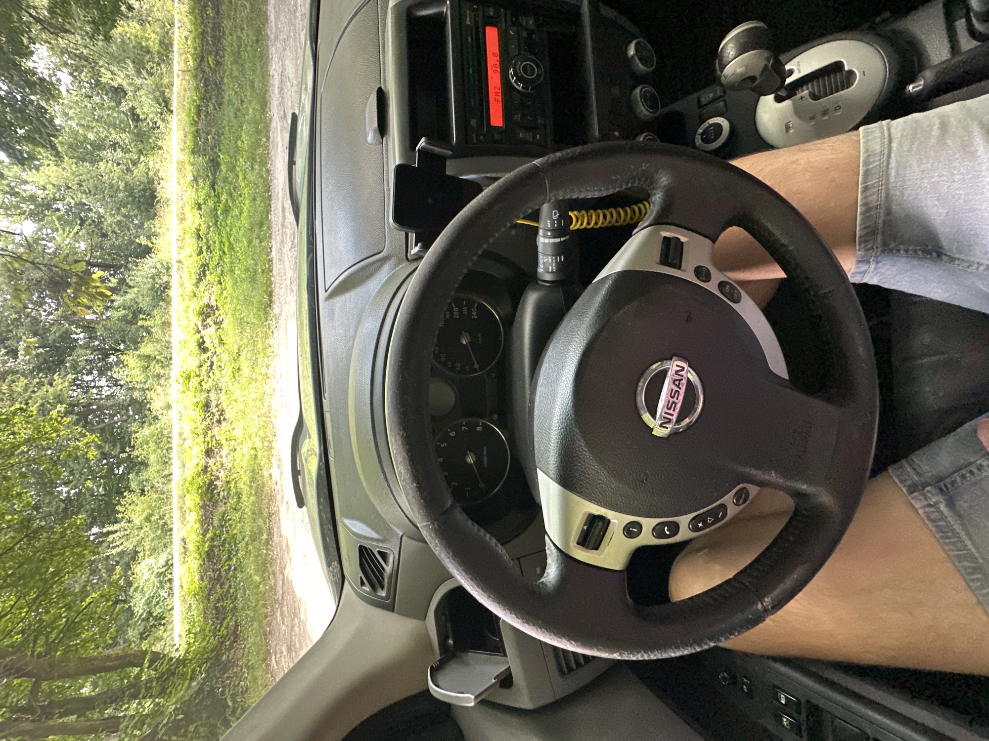 The image, taken from inside a Nissan vehicle, shows a view of the driver's dashboard, steering wheel, and the legs of the driver wearing denim shorts. Outside the car, lush green trees and vegetation are visible, suggesting a rural or natural setting. The car is stopped, possibly on a dirt or gravel road. The radio display indicates "FM2 98.8". The location is Shatava, Ukraine.