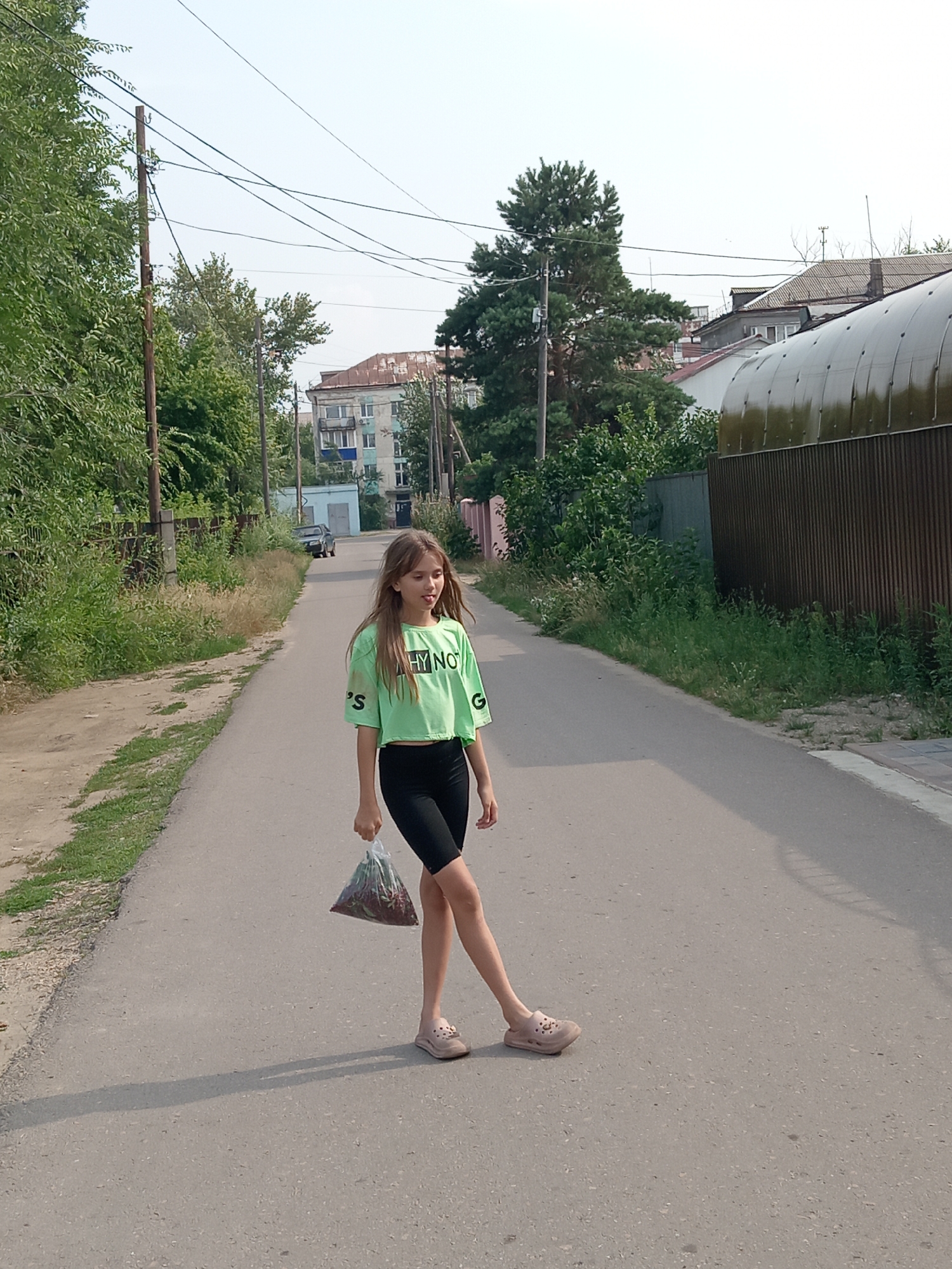 In Rudny, Kazakhstan, a young girl with long, light brown hair walks down a paved road. She is wearing a light green, cropped t-shirt with lettering and black athletic shorts. In her right hand, she holds a clear plastic bag filled with what appears to be red berries. Her left leg is extended forward, and she is looking to her right. The road is bordered by overgrown green grass and foliage on the left, and a tall, corrugated metal fence and a building on the right. Overhead, utility wires stretch across the sky. Residential buildings and trees are visible in the background, suggesting a semi-urban or suburban setting. The lighting indicates it is daytime, with a slight haze in the sky.