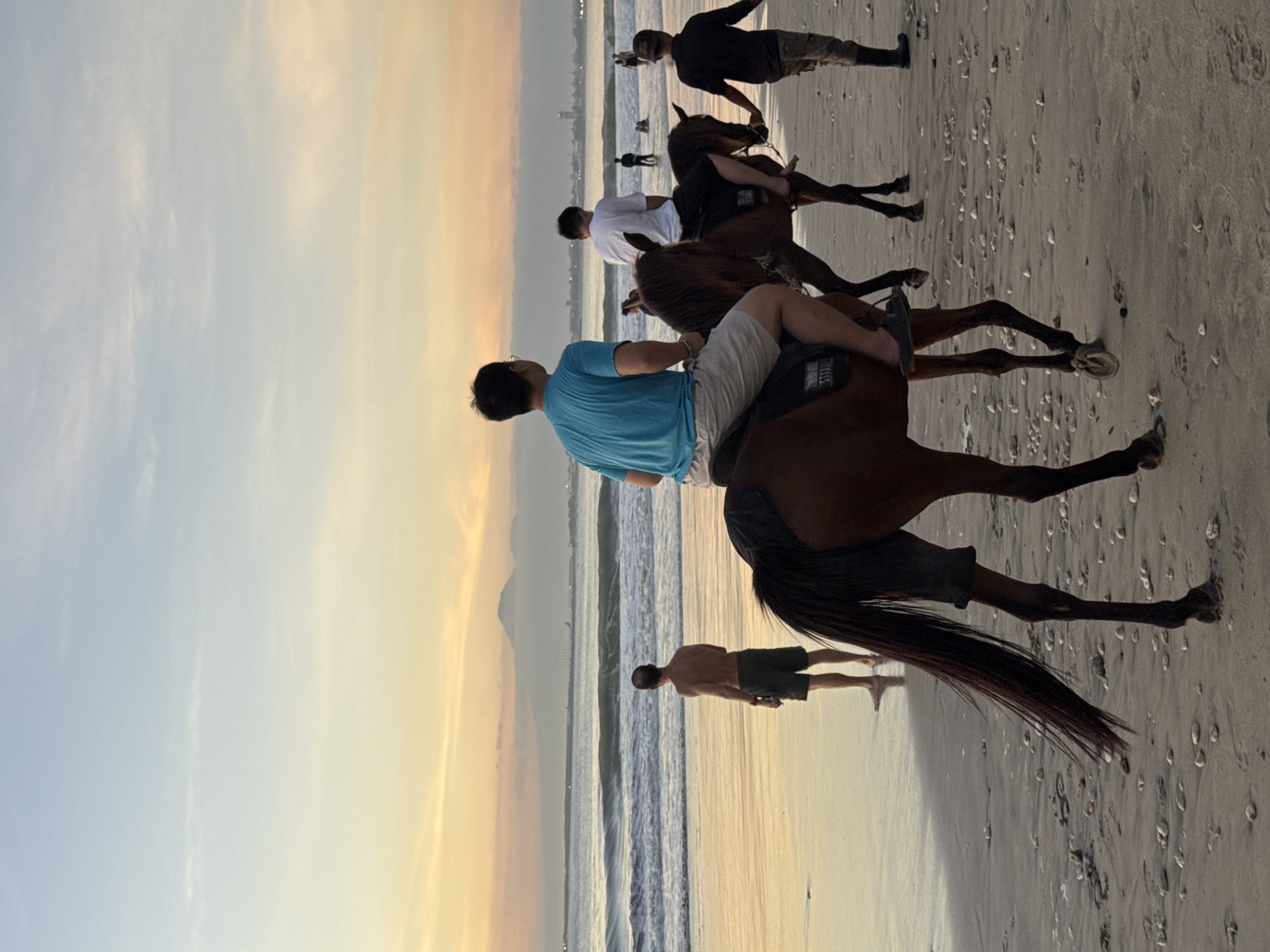 The image shows a beach scene in Kuta Selatan, Indonesia. Three brown horses are being ridden along the beach. The riders are all facing the same direction, toward the right of the frame. The first rider is wearing a blue shirt and light-colored shorts. The second rider is wearing a white shirt. The third rider is wearing a black shirt and light-colored pants. A man is walking on the beach near the water. The sky is a mix of yellow and blue, suggesting it is either sunrise or sunset. The beach is sandy with scattered shells and footprints. The ocean waves are visible in the background.
