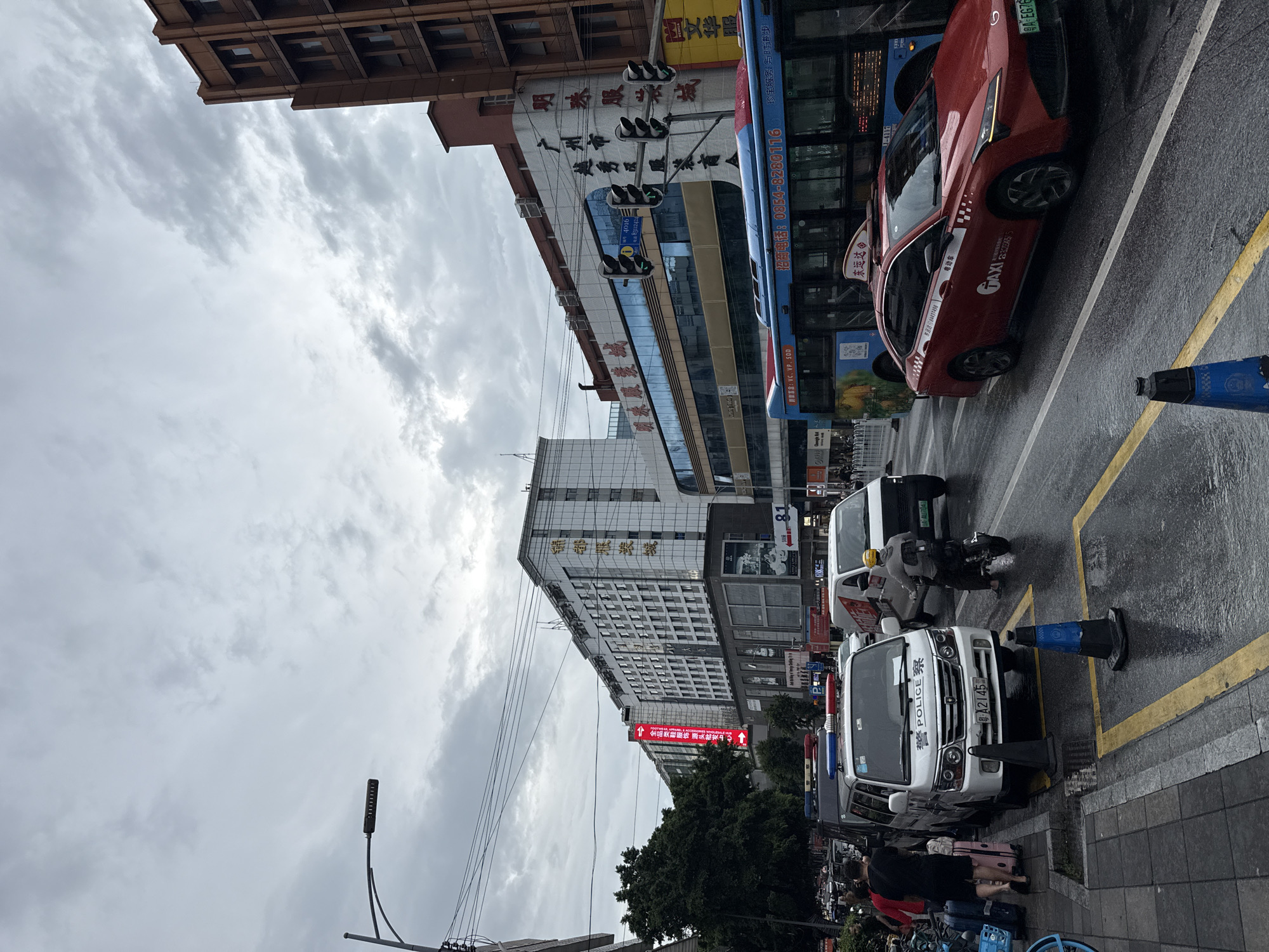 The image depicts a street scene in Yuexiu District, China, under an overcast sky. In the foreground, a wet asphalt road is visible with yellow painted lines and traffic cones. A red taxi and a police van are parked on the side of the road. The police van has "POLICE" written on it in English and Chinese characters, and a police light bar is on its roof. Several people are on the sidewalk, including one person walking with luggage. Further down the street, a blue bus is driving, and various buildings line the road, some with prominent signage in Chinese. The buildings appear to be commercial establishments, with a mix of modern and older architecture. Traffic lights and electrical wires are also visible. The overall impression is a busy urban environment on a cloudy day.