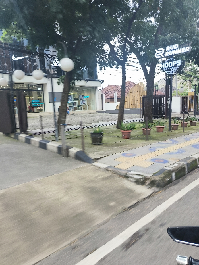 The image shows a street in Bandung, Indonesia, located at the coordinates -6.899, 107.621. A black and white checkered curb borders the street, which is paved with grey asphalt. A white line separates the street from a small, grassy area. The grassy area has several potted plants in it, including one in the shape of a basketball. Two large trees are in the background and a large building with a storefront and a storefront with a sign that reads "RUD RUNNER HOOPS". The storefront is open and there is a person standing in the background. The image was captured from a moving vehicle, and the street is blurred. The scene is well lit and the weather is clear.